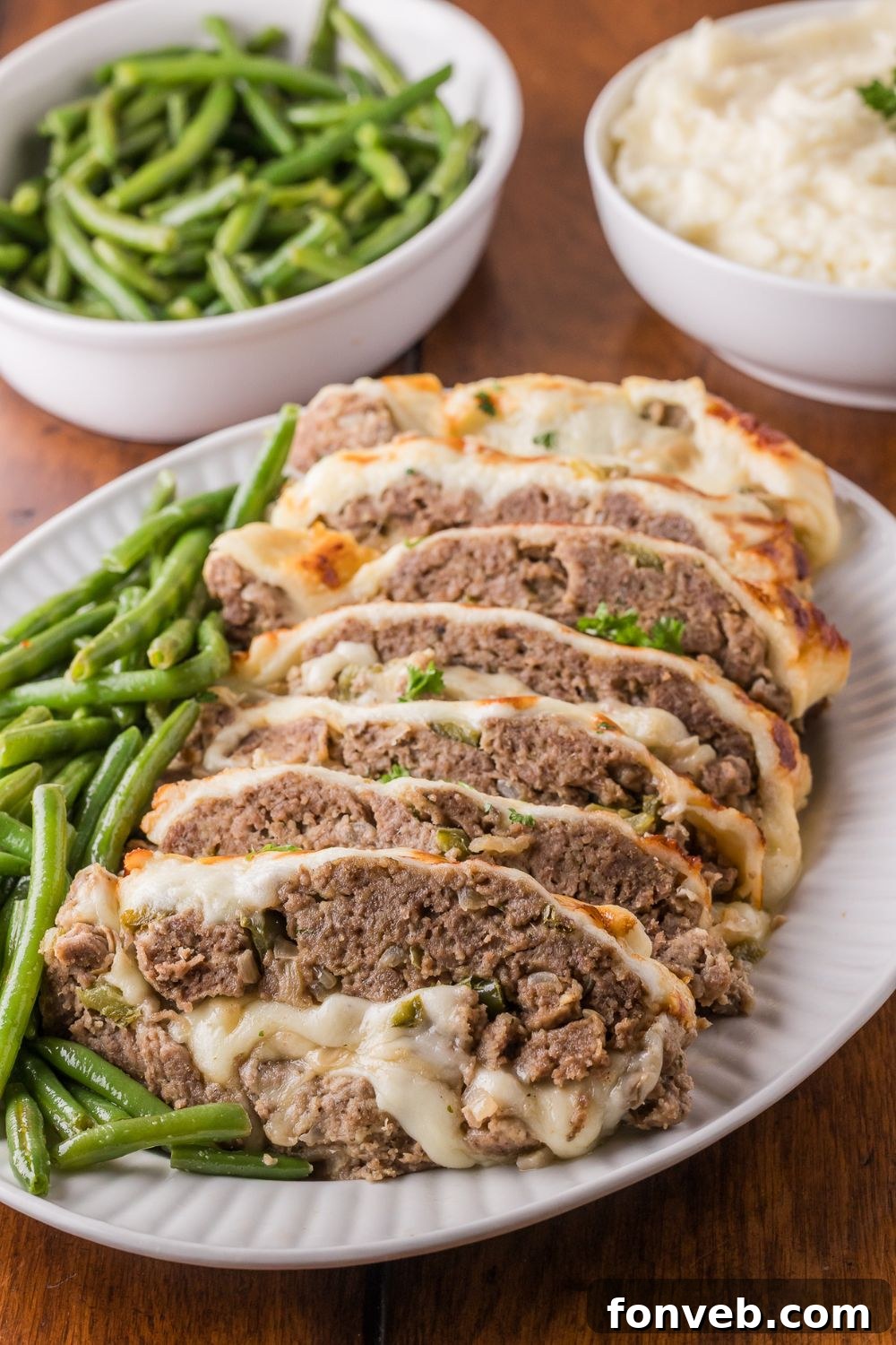 wooden table with a bowl of green beans, bowl of mashed potatoes, and a platter with sliced Stuffed Philly Cheesesteak Meatloaf on platter with green beans on side