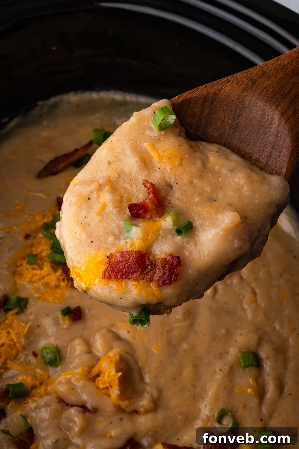 wooden bowl with some of the loaded potato soup with cheese, bacon and green onions in a black crockpot