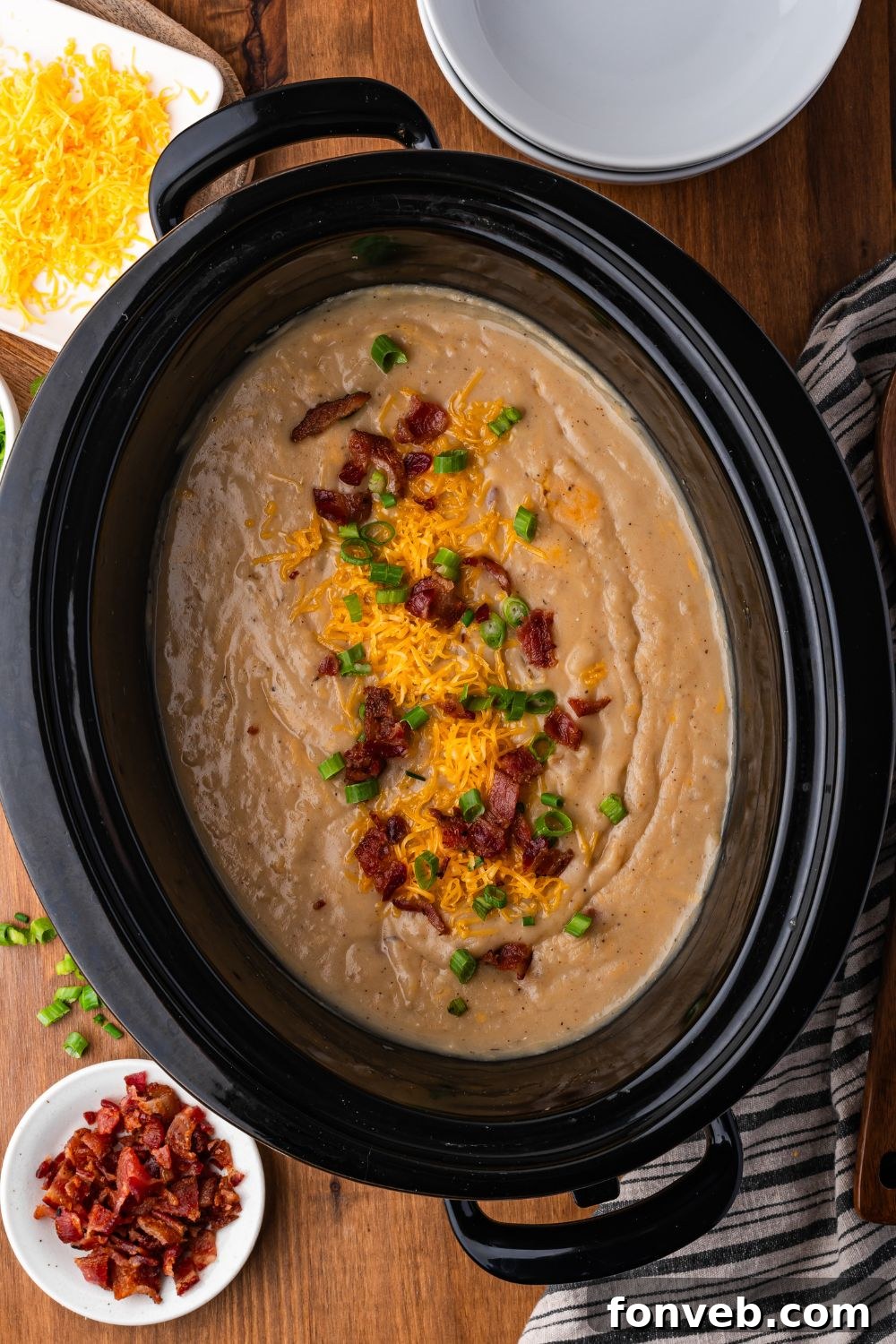 wooden table with bowls of shredded cheese, chopped bacon, and more in white bowls. Then in center is a black crockpot full of creamy loaded baked potato soup. It is topped with crumbled bacon, green onions and shredded cheese