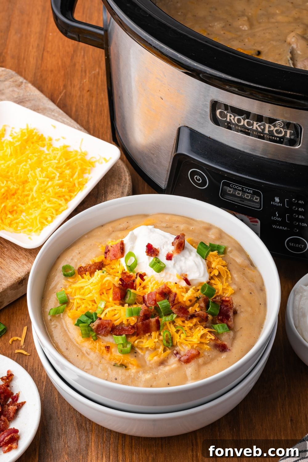 Crockpot in the background of picture. A wooden table that has two white bowls stacked and the top bowl is full of potato soup topped with shredded cheese, bacon, onions and a dollop of sour cream