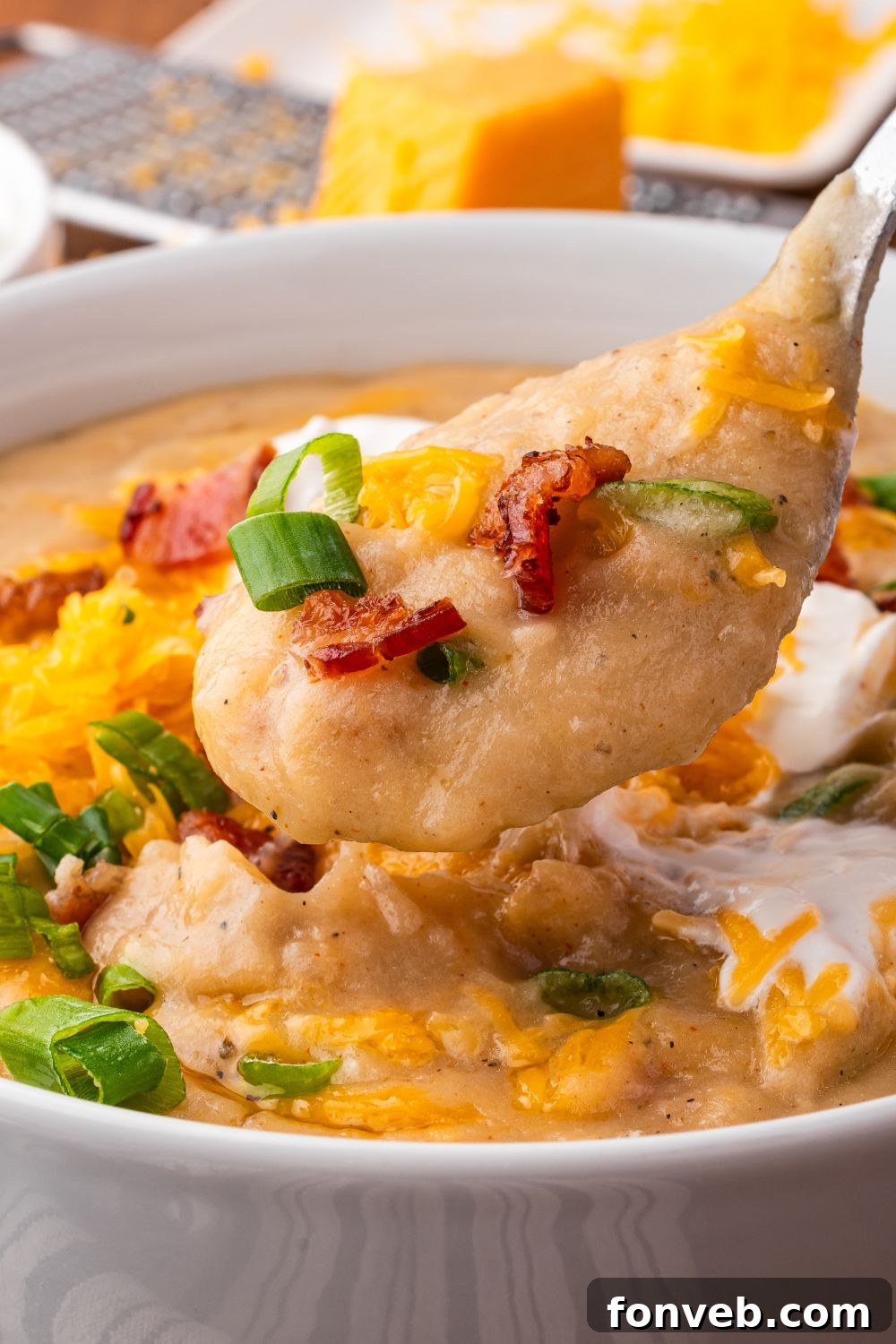 close up of white bowl on table with Crockpot Loaded Baked Potato Soup in it, and topped with crumbled bacon, green onion and shredded cheese. In  the background there is a cheese grater with a block of cheese 
