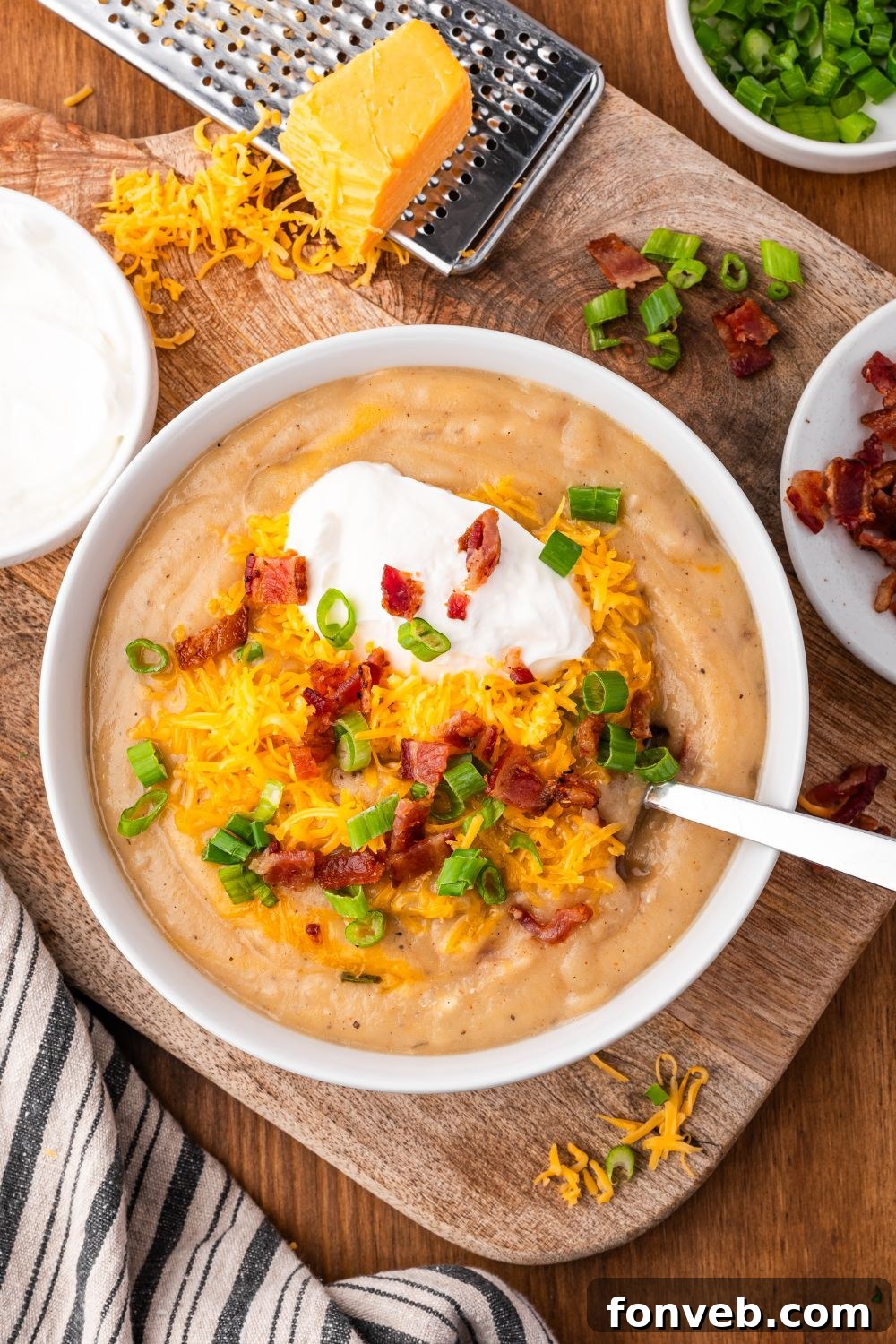 wooden table with a cheese grater with a block of cheese on top. A white bowl full of Crockpot Loaded Baked Potato Soup with toppings and a spoon to side. And crumbles of cheese around the bowl 