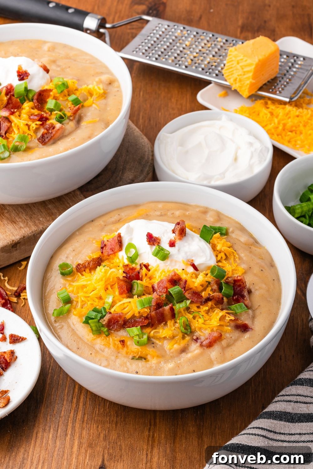 white bowls on table with Crockpot Loaded Baked Potato Soup in each bowl. They are topped with soup toppings and around the wooden table are small bowls of green onions, sour cream, shredded cheese, etc. 