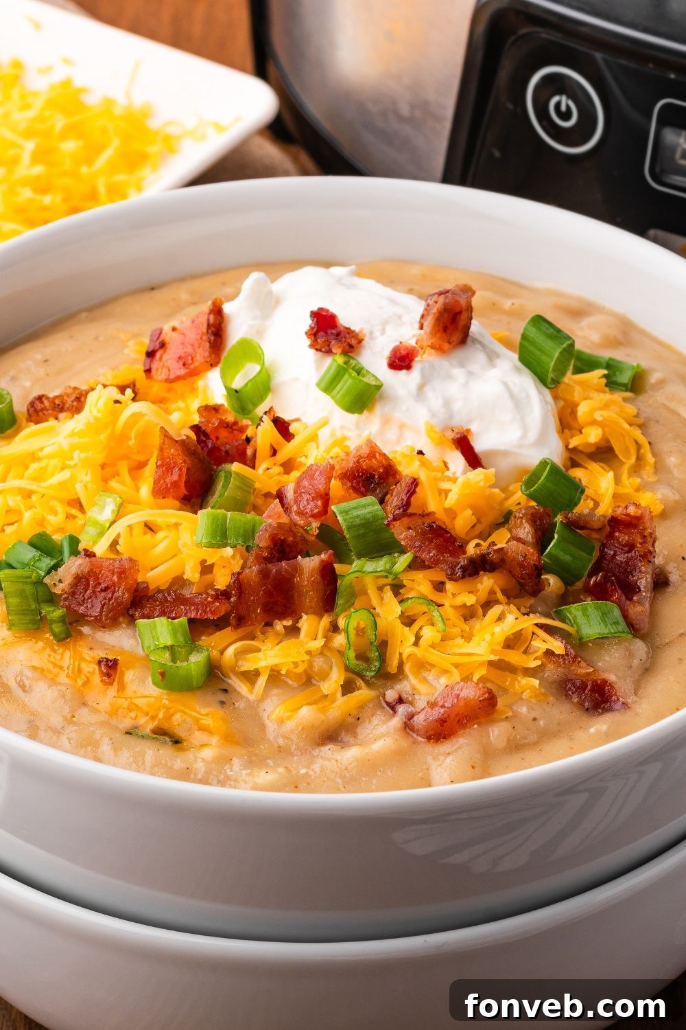 two white bowls stacked and top one full of Crockpot Loaded Baked Potato Soup with toppings . Behind the bowls is a silver and black crockpot and a small bowl of shredded cheese. 