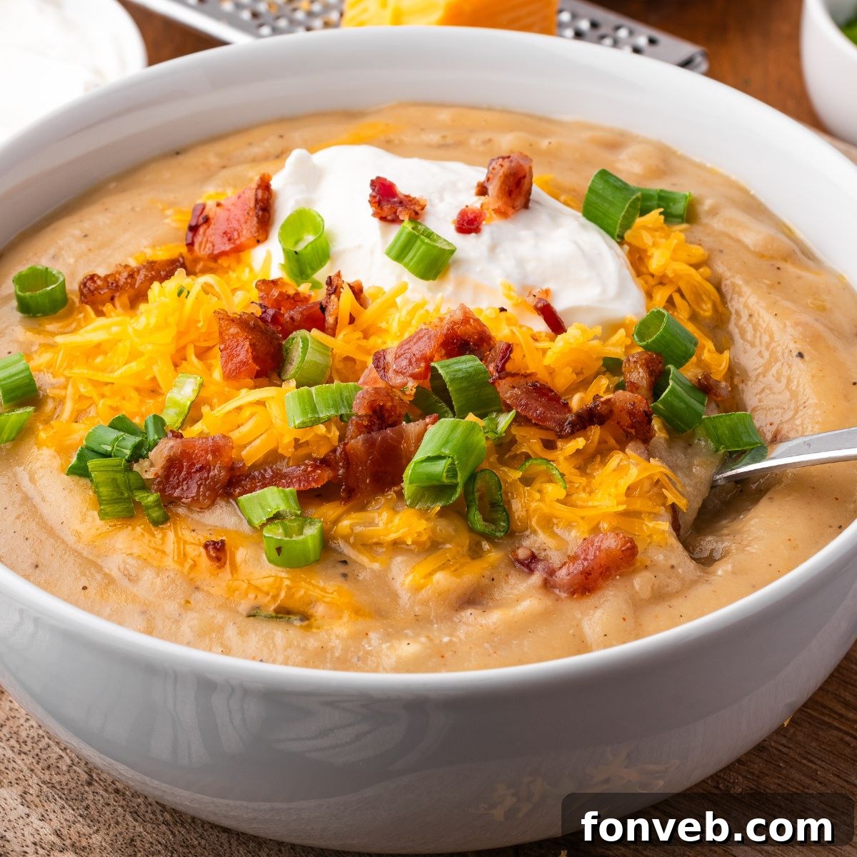 wooden table with a white bowl full of Crockpot Loaded Baked Potato Soup topped with shredded cheese, bacon crumbles, green onions and sour cream