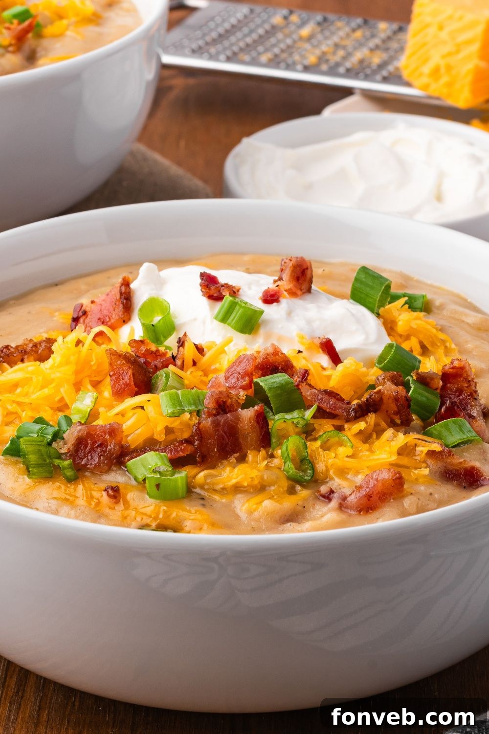 side view of table with two stacks of white bowls full of Crockpot Loaded Baked Potato Soup topped with shredded cheese, green onions and back bits and sour cream. Behind the table is a bowl of sour cream 
