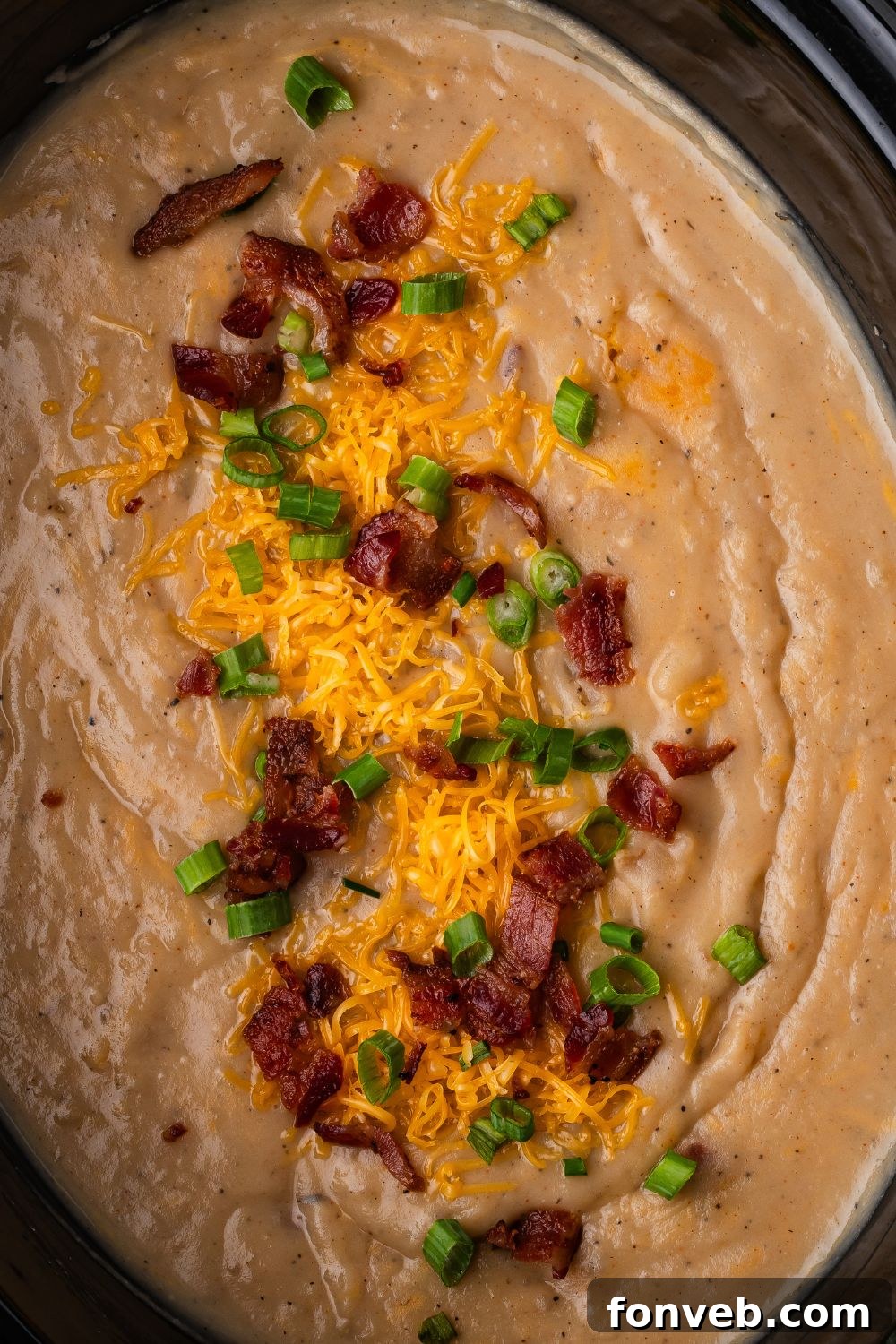 a black crockpot sitting on table that is full of loaded baked potato soup. Then on top you have shredded cheese, bacon, and chopped green onions