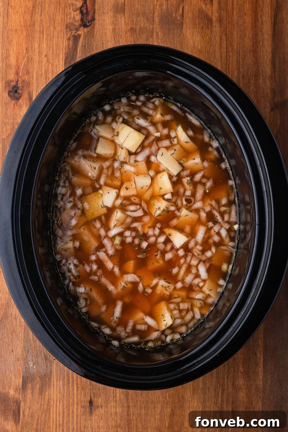 black crockpot sitting on a wooden  table with diced potatoes, onions, beef broth in more in the slow cooker