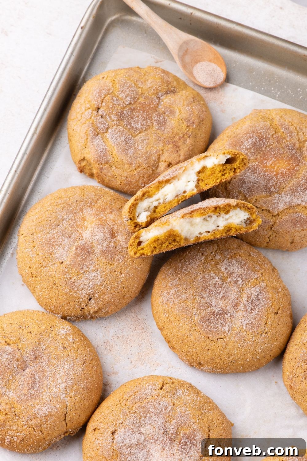 sheet pan on white table with pumpkin cookies on sheet with one cookie broken in half to see the cheesecake filling 