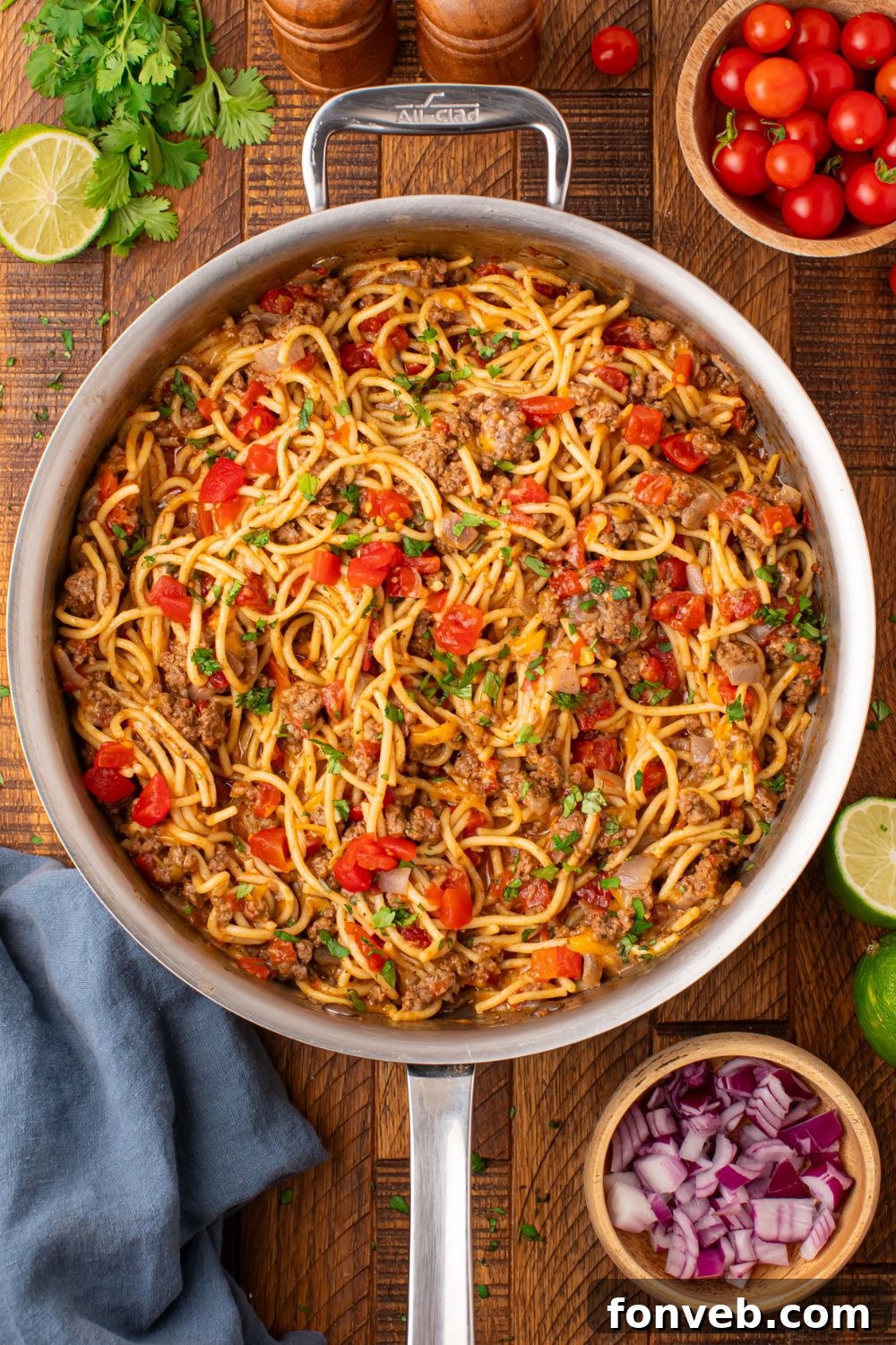 One Pot Cheesy Taco Spaghetti 13 looking down on wooden table with a large metal skillet with taco spaghetti. Then cilantro, tomatoes, lime and more around table