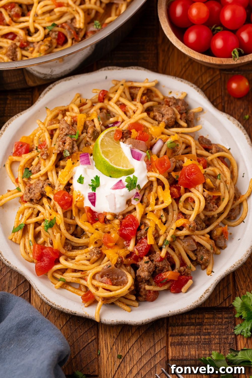 One Pot Cheesy Taco Spaghetti 4 Table set up for serving up dinner with a bowl of tomatoes to side, skillet with more pasta, and a large white place and topped with sour cream, and a lime wedge