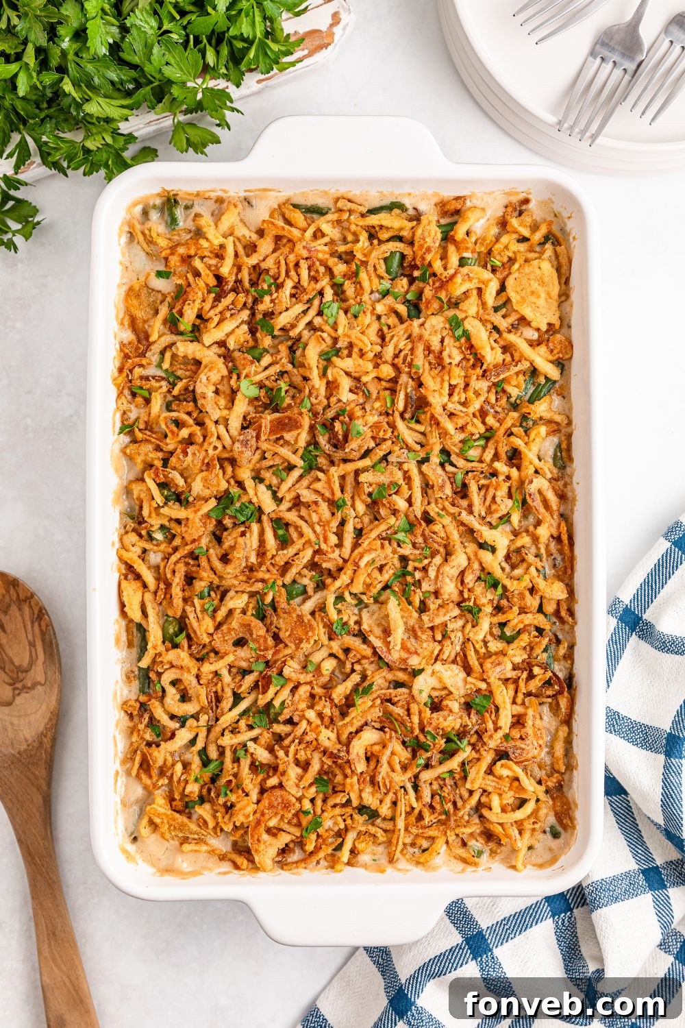 Green Bean Casserole in a white dish on table with a wooden spoon, towel and more placed around pan 