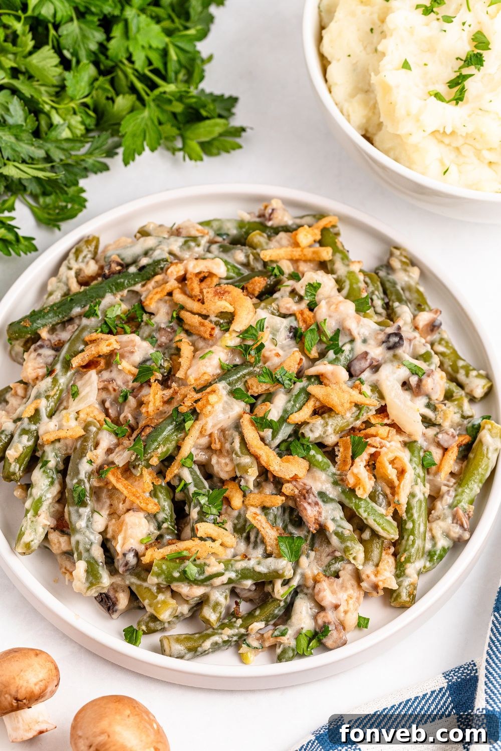 Christmas green bean casserole in a round white dish on table with mashed potatoes in a bowl to side and fresh herbs