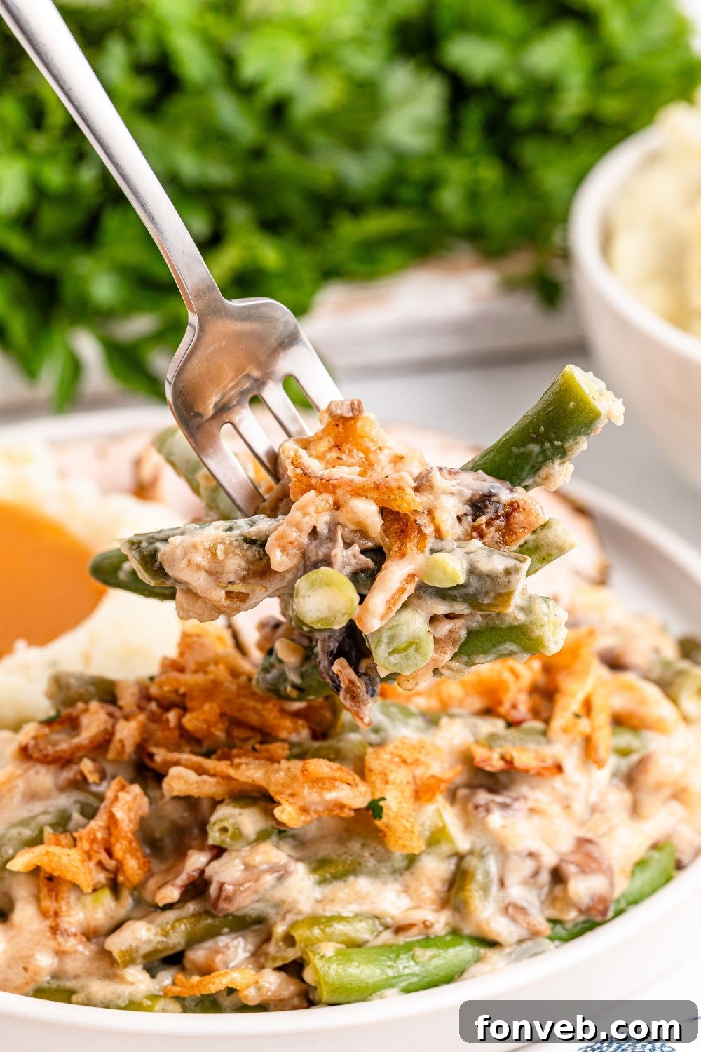Green Bean Casserole on a white plate on table with a fork scooping up some of the mixture. Parsley greens are behind it on table 