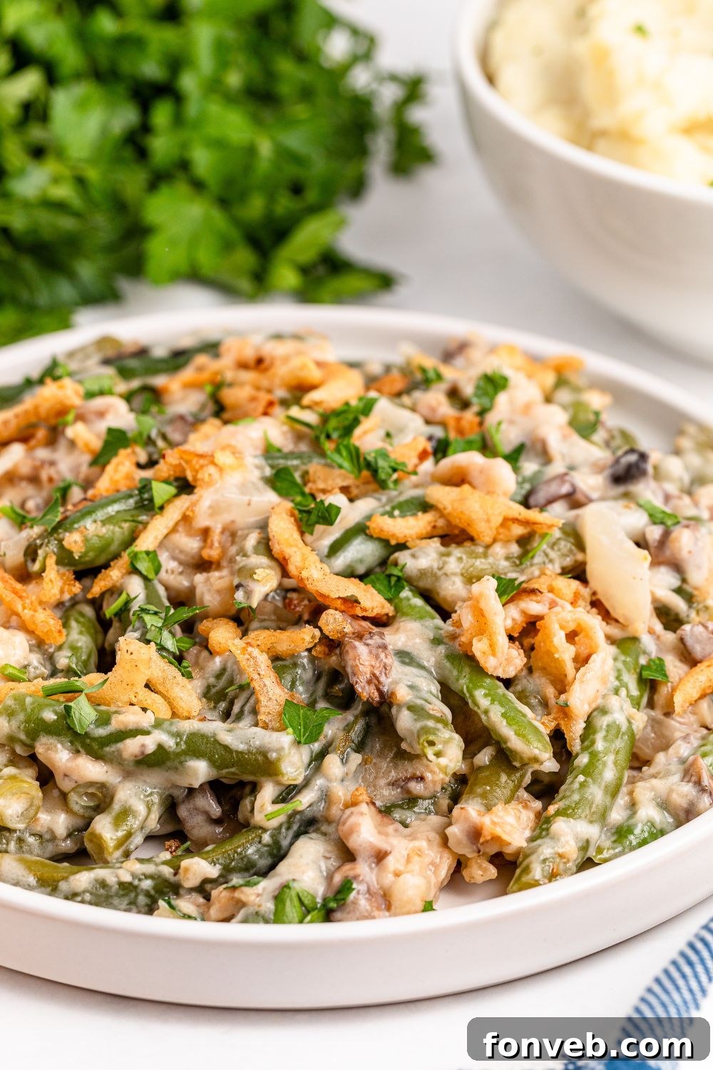 side view of a white plate with an edge sitting on table full of Green Bean Casserole and behind it on table fresh parsley bundle 