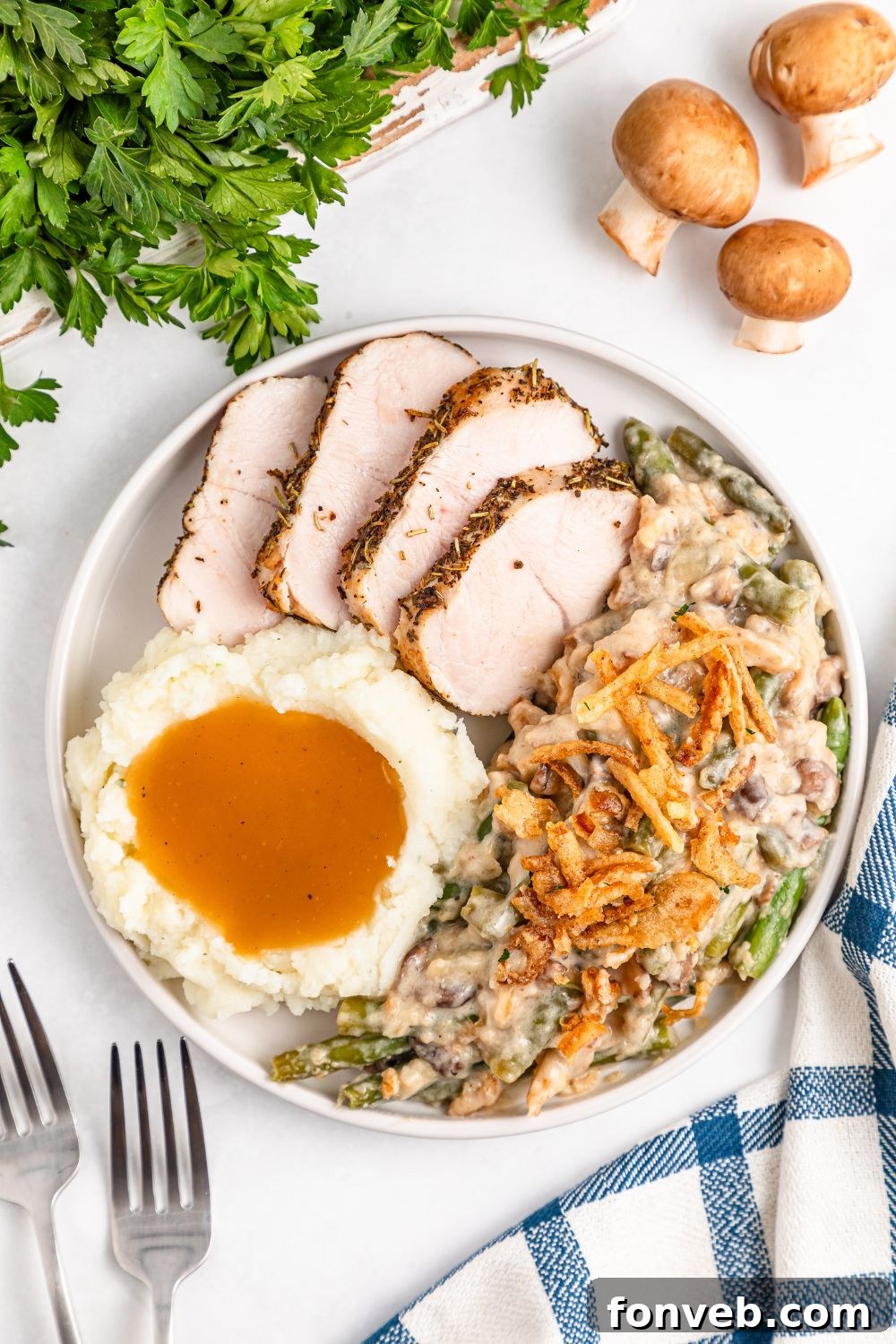 looking down on a white table with a plate full of holiday food- sliced turkey, mashed potatoes, gravy, and green bean casserole. Mushrooms and parsley are around table for decor 