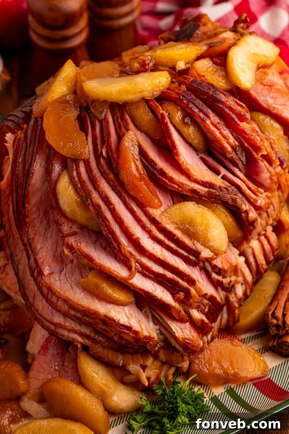 Tender and Juicy Slow Cooker Apple Glazed Ham 18 view of a large sliced spiral ham on platter with some apples on top and around with a red checkered towel behind it
