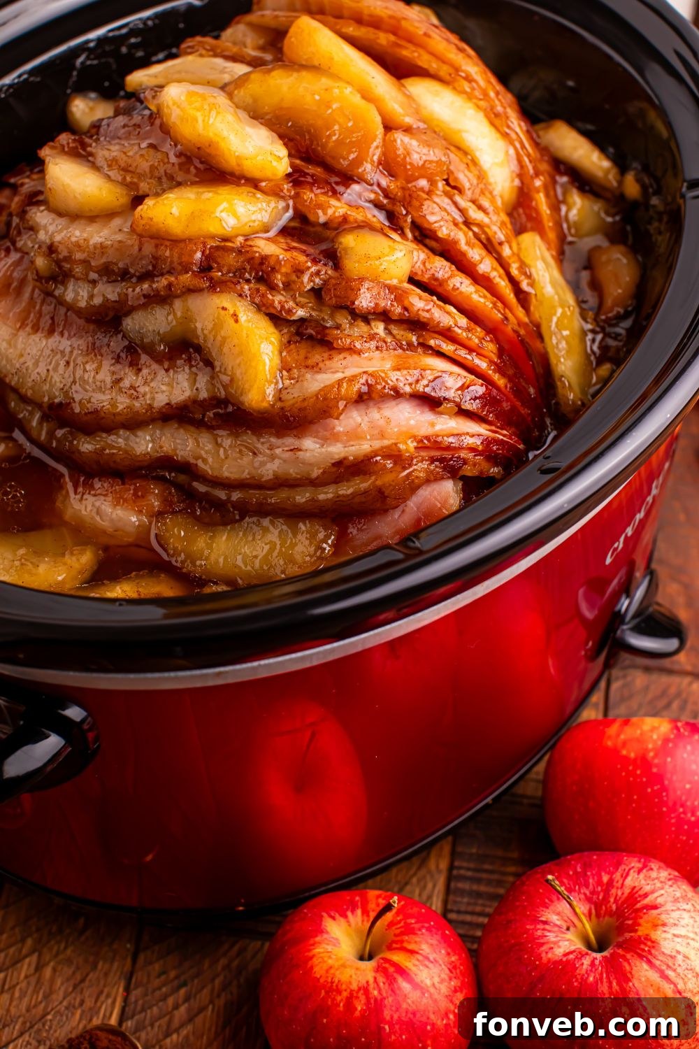Tender and Juicy Slow Cooker Apple Glazed Ham 9 red slow cooker on a wooden table with a spiral ham with apples on top, and whole apples in front of table