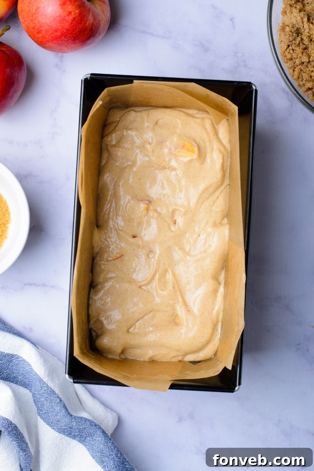 Quick Glazed Apple Cinnamon Loaf 11 A bird's-eye view of a marble counter featuring a loaf pan partially filled with a smooth cinnamon loaf cake batter, with fresh apples and a crumbly brown sugar mixture positioned beside it.