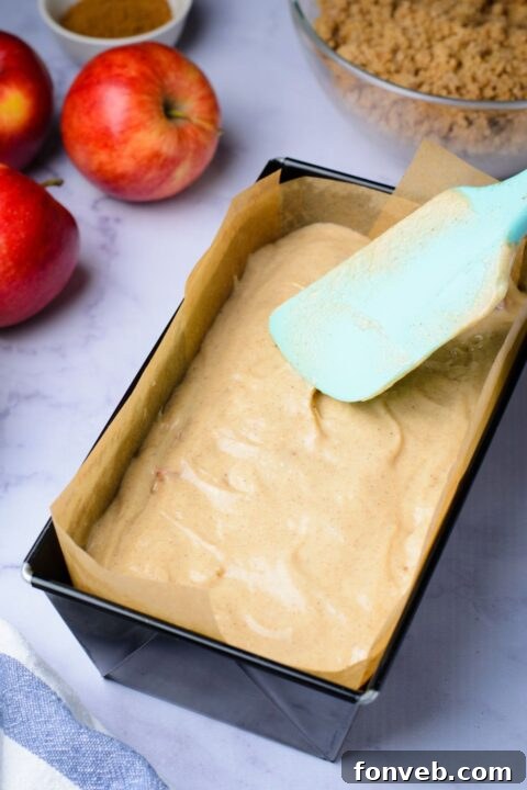 Quick Glazed Apple Cinnamon Loaf 13 Raw batter in a loaf pan is being gently spread with a spatula, creating an even layer for baking, with two fresh apples resting on the table beside the pan.