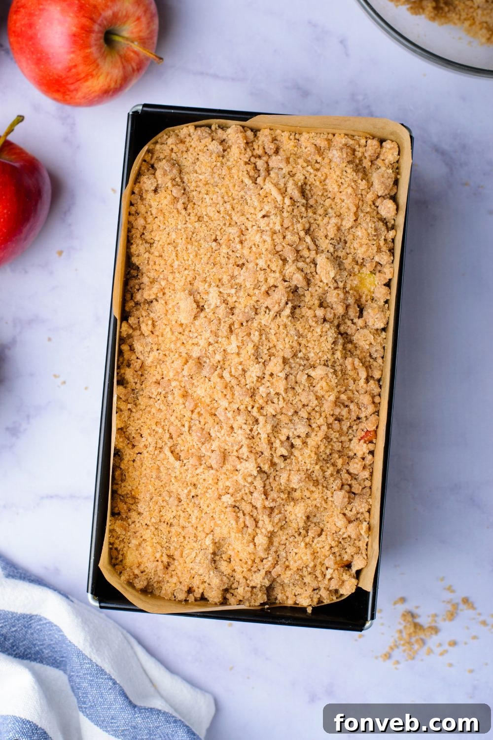 Quick Glazed Apple Cinnamon Loaf 14 A rich cinnamon crumble topping is artfully spread over the raw loaf cake batter in the pan, with a couple of fresh apples scattered nearby on the table, indicating the final prep stage.
