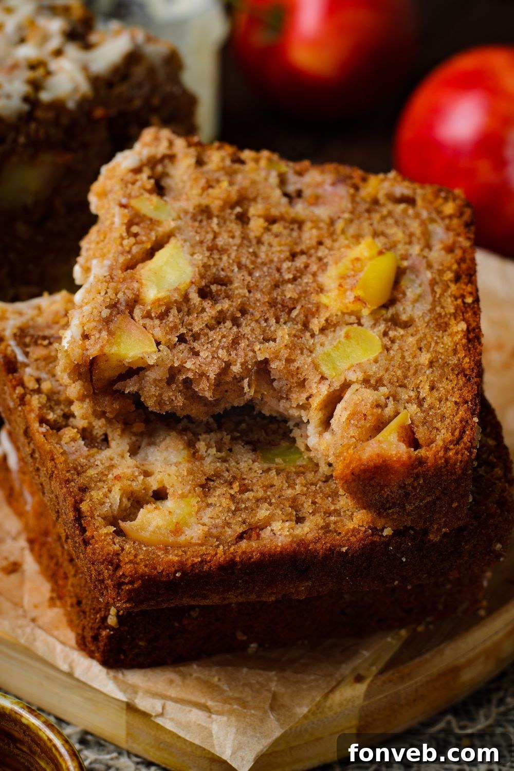 Quick Glazed Apple Cinnamon Loaf 17 Thick, glazed slices of Apple Cinnamon Loaf Cake are stacked elegantly on a tray, with one slice showing a bite taken out, complemented by vibrant red apples in the background for a rich autumn aesthetic.