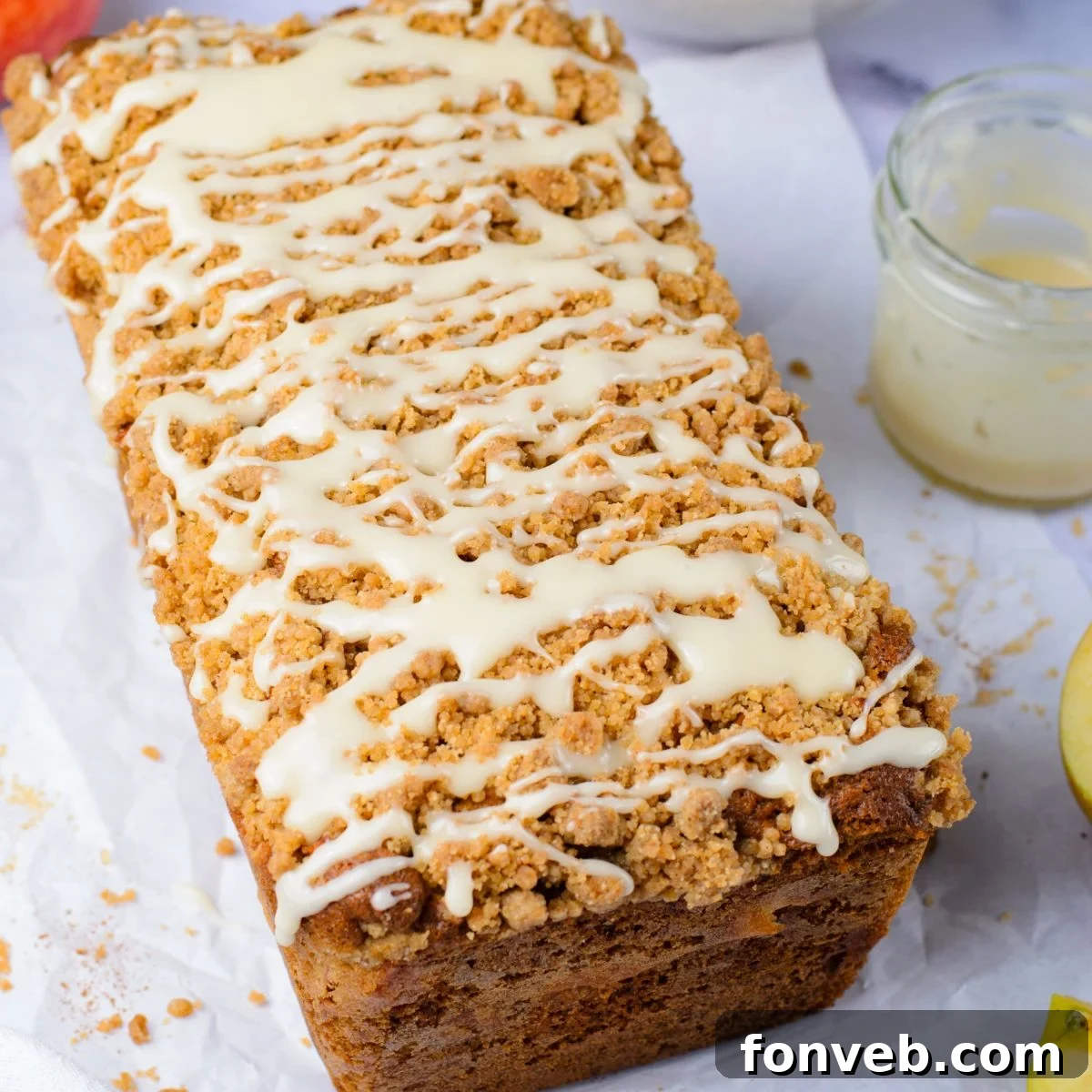 Quick Glazed Apple Cinnamon Loaf 3 A freshly baked Apple Cinnamon Loaf Cake, drizzled with white glaze, resting on a wooden table alongside a jar of extra glaze and fresh apples, evoking a warm autumn scene.