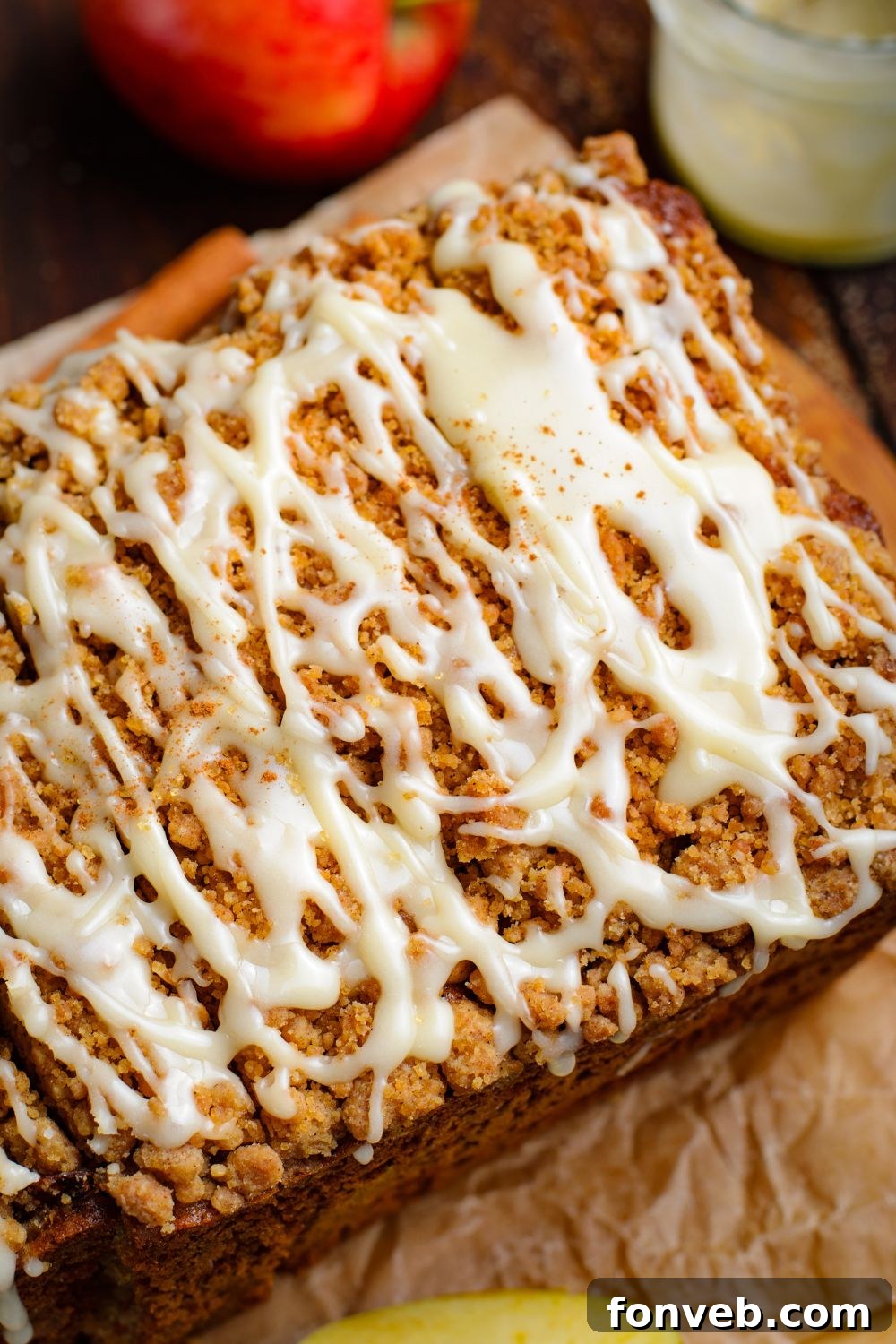 Quick Glazed Apple Cinnamon Loaf 28 An Apple Cinnamon Loaf Cake sits elegantly on parchment paper on a wooden table, with a glass of sweet glaze and a vibrant red apple positioned to the side, creating a rustic and inviting culinary scene.