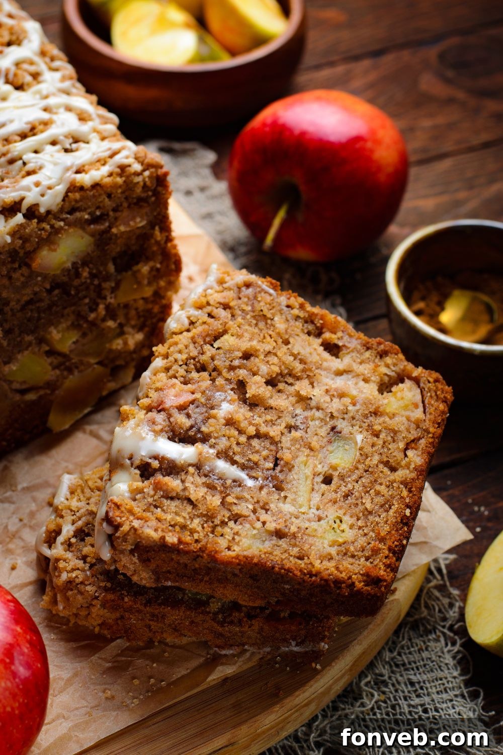 Quick Glazed Apple Cinnamon Loaf 29 A delicious Apple Cinnamon Loaf Cake sits on a wooden table, with some of its thick slices neatly stacked. A wooden bowl of sliced apples and a whole apple are arranged decoratively to the side, enhancing the autumnal theme.