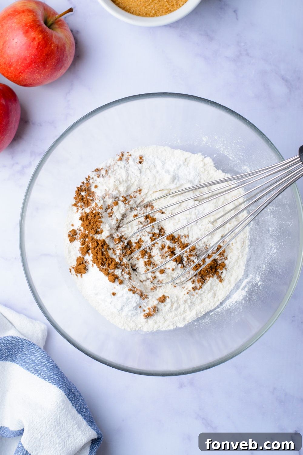 Quick Glazed Apple Cinnamon Loaf 7 A close-up of dry ingredients, flour and cinnamon mixture, in a clear glass bowl on a marble counter, with two apples positioned decoratively to the side.