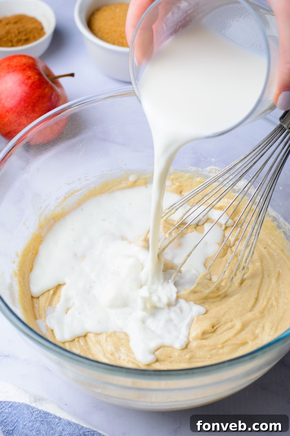 Quick Glazed Apple Cinnamon Loaf 9 A vibrant red apple rests beside a glass bowl containing wet batter, with buttermilk being poured in and a whisk lying nearby, indicating the mixing process for the loaf cake.