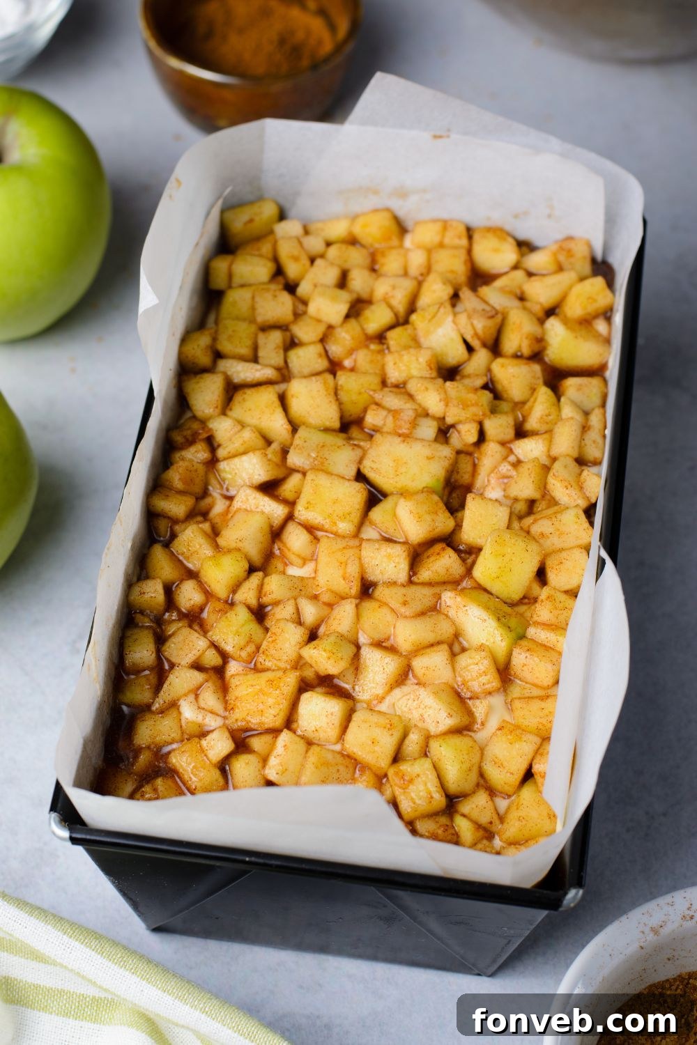 diced apples in a loaf pan for making a fritter bread with apples around the pan on table and a cinnamon mixture in a bowl