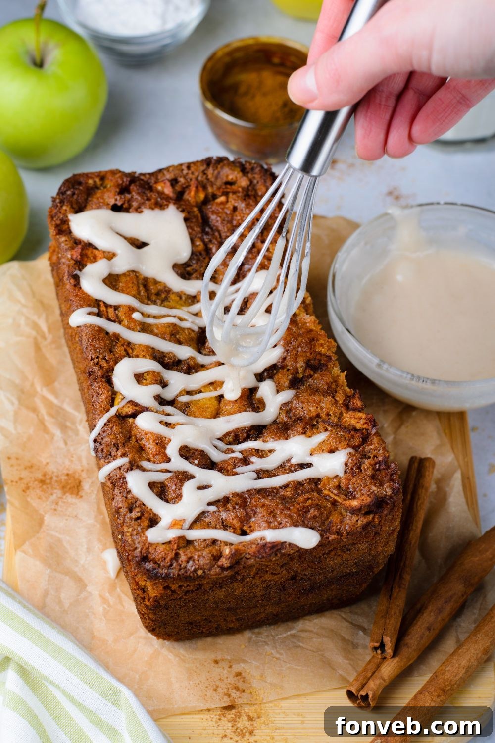amish apple bread on table with a whisk drizzling glaze over the top with a small bowl of glaze to side 