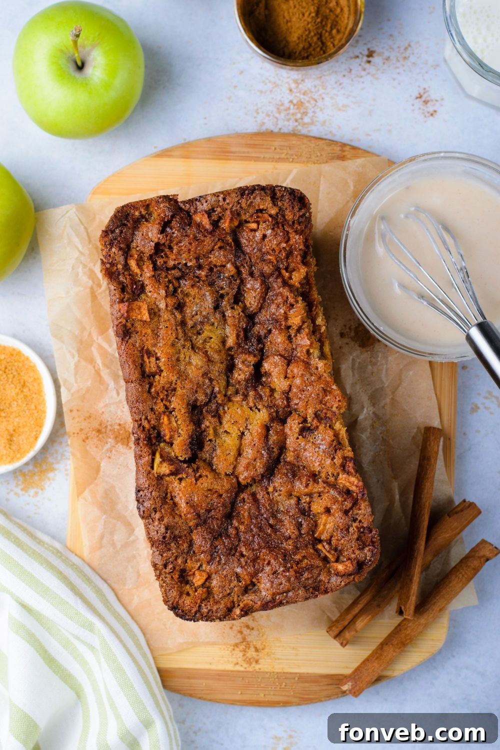 apple fritter bread sitting on marble counter top with glaze in a bowl to side, cinnamon sticks, apples and more scattered around table 