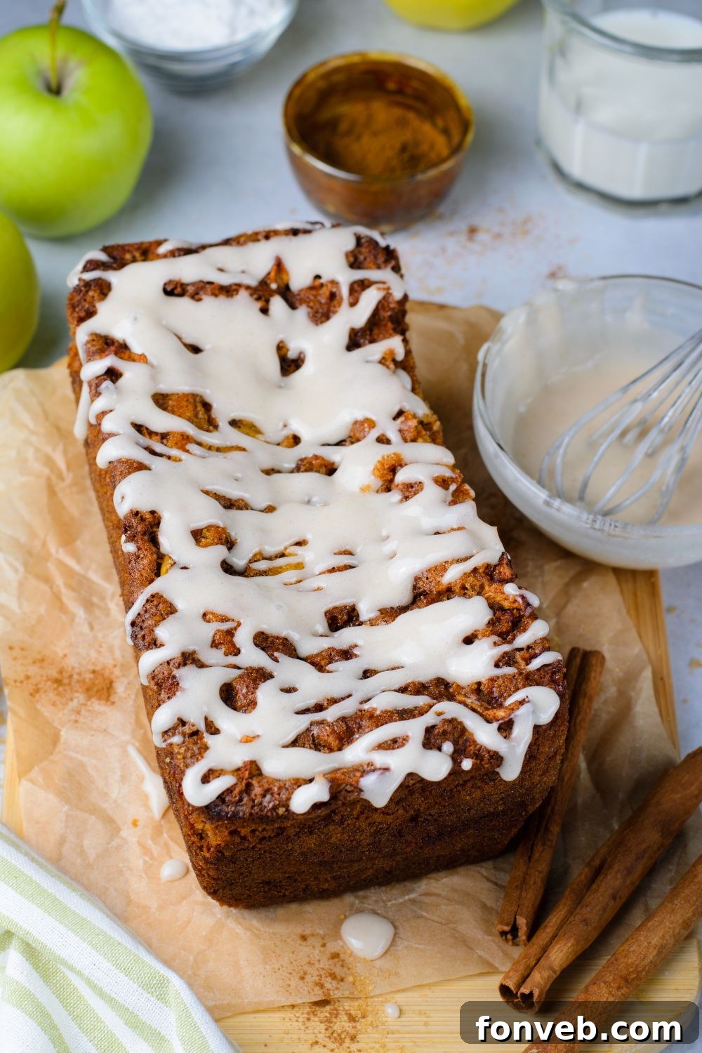 Amish Apple Fritter Bread on brown parchment paper on table with a bowl of glaze to side and cinnamon sticks in front of loaf 