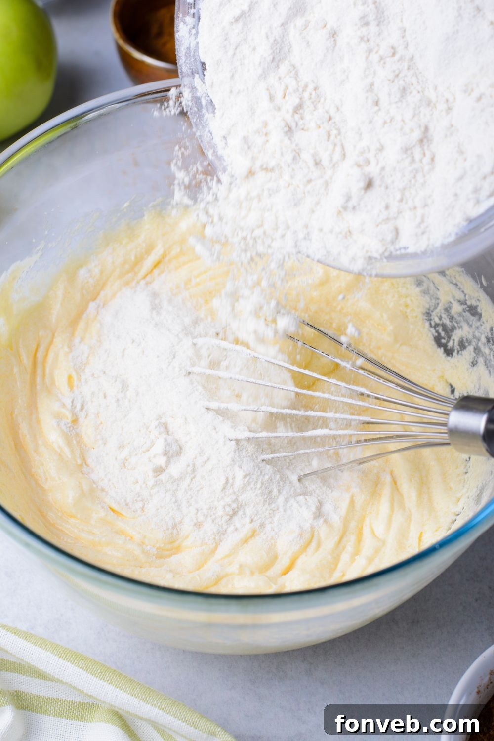 a bowl pouring flour mixture into a glass bowl of wet batter for making Amish Apple Fritter Bread