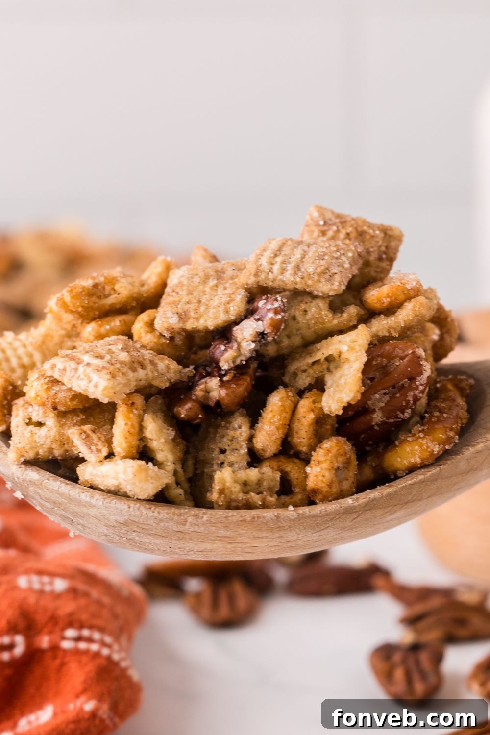 Pumpkin Spice Chex Mix in a wooden spoon with some nuts and more snack mix on the counter with an orange towel in front of table 