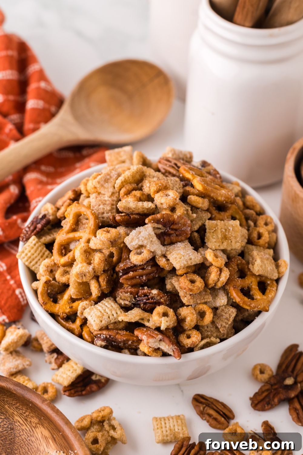 orange towel laid out on table with a wooden spoon in the back, and a bowl full of Pumpkin Spice Chex Mix and more snack mix spread around the table 