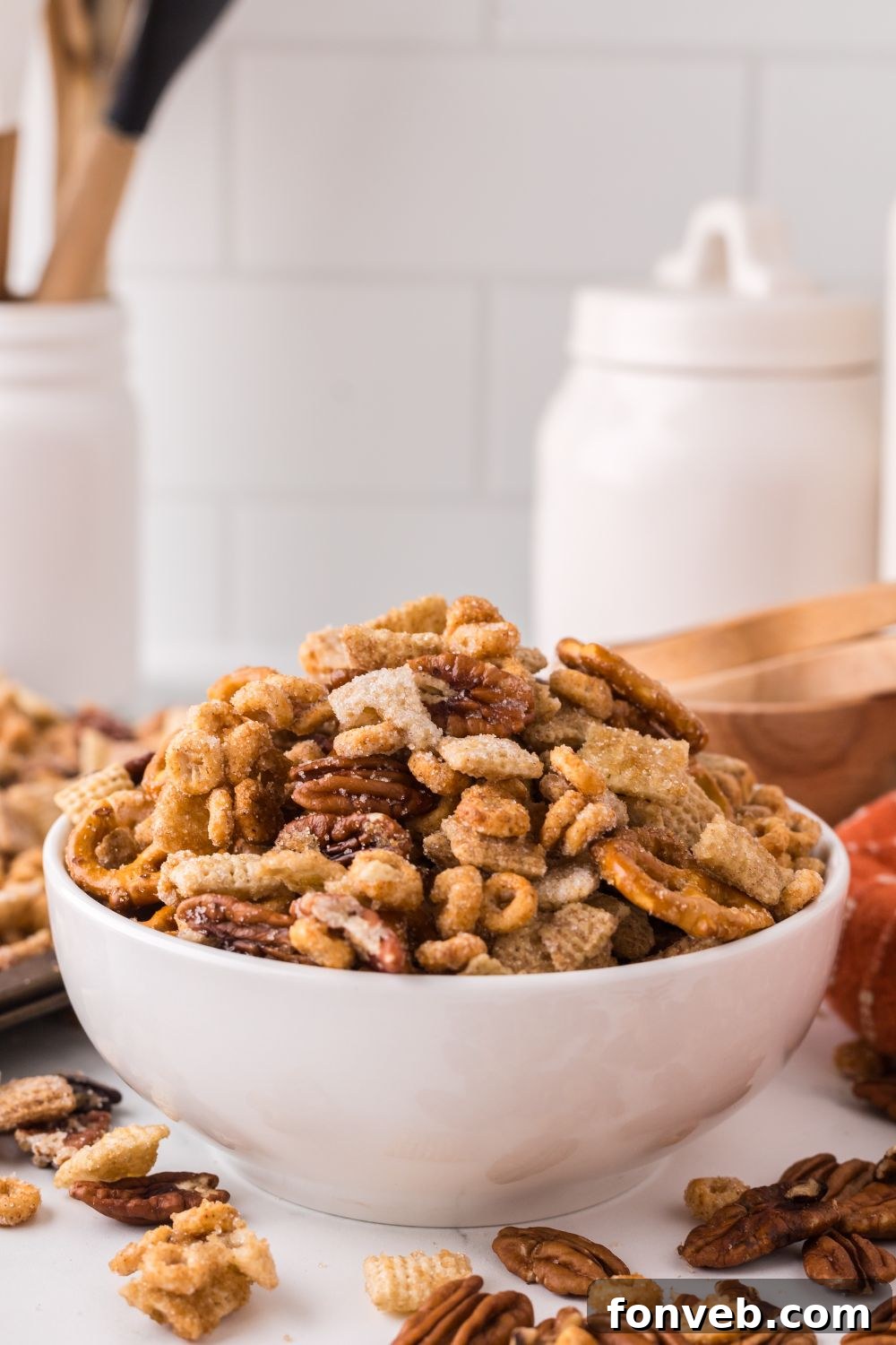 white kitchen counter that has a wooden cutting board in back, and in front is a white bowl with Pumpkin Spice Chex Mix inside, and more nuts and cereal mix spread out around the table 
