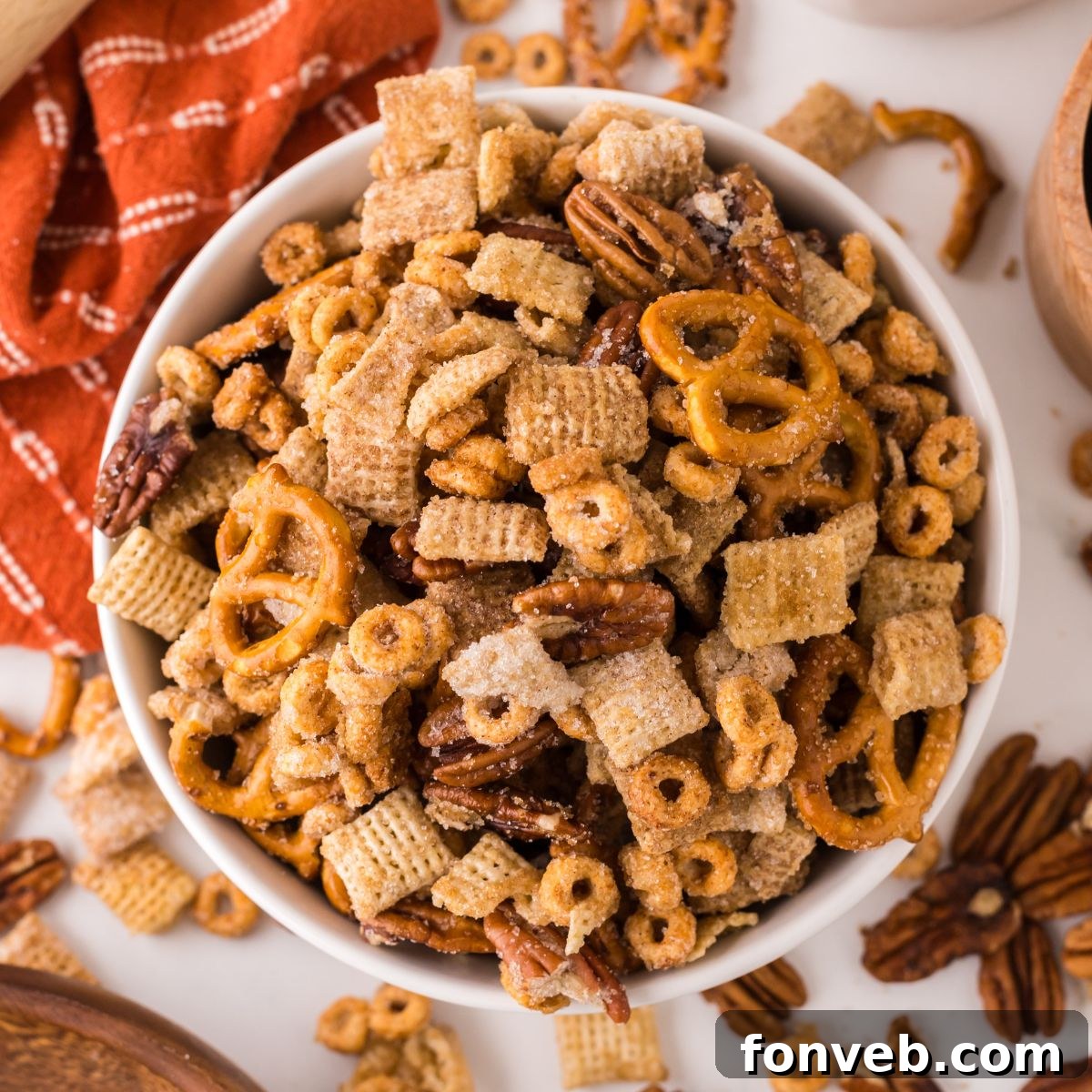 Pumpkin Spice Chex Mix in a white bowl on table with more mix scattered on table with a towel to the side of the bowl 
