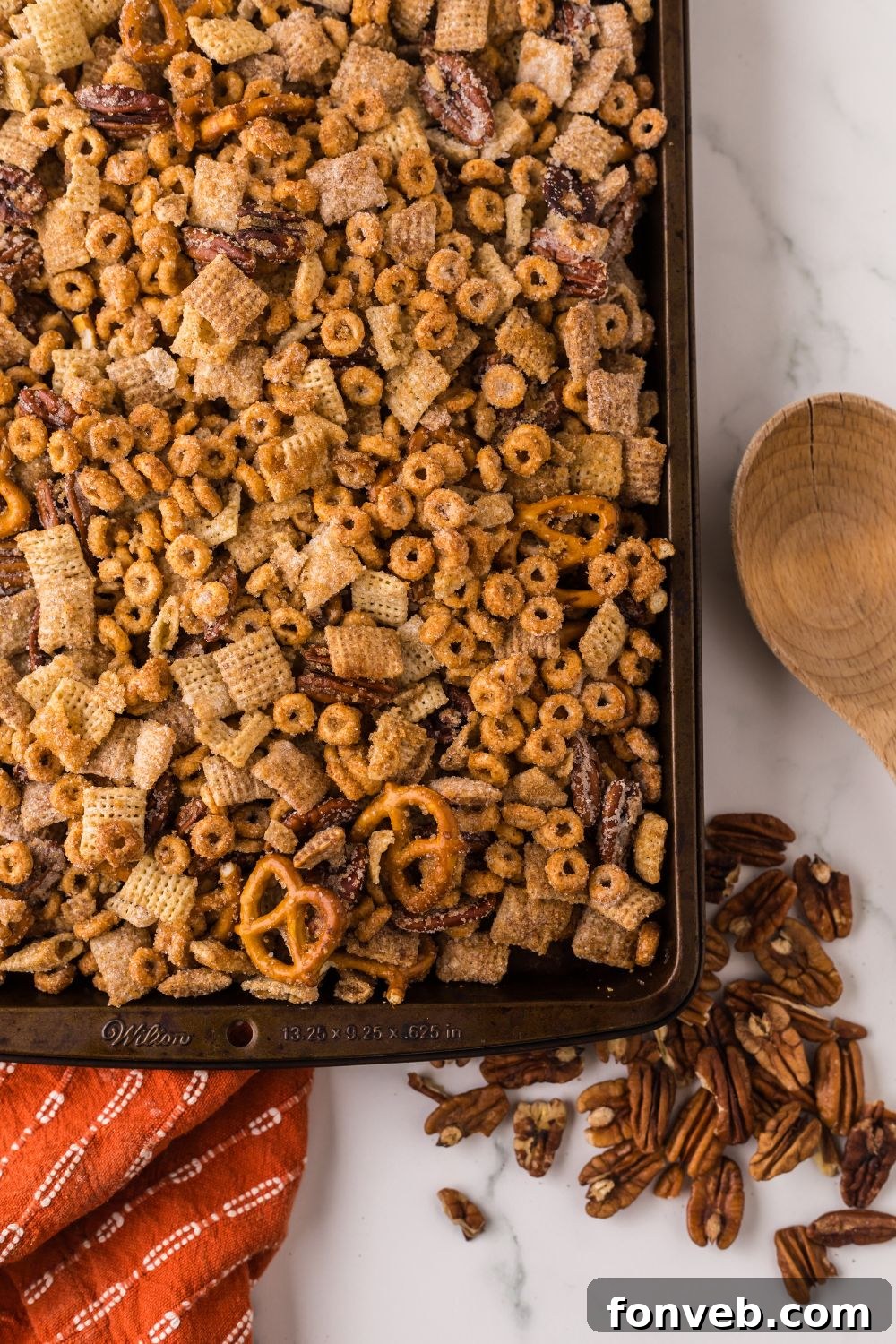 looking down at counter with a sheet pan full of Pumpkin Spice Chex Mix and some nuts scattered on counter with a wooden spoon to the side