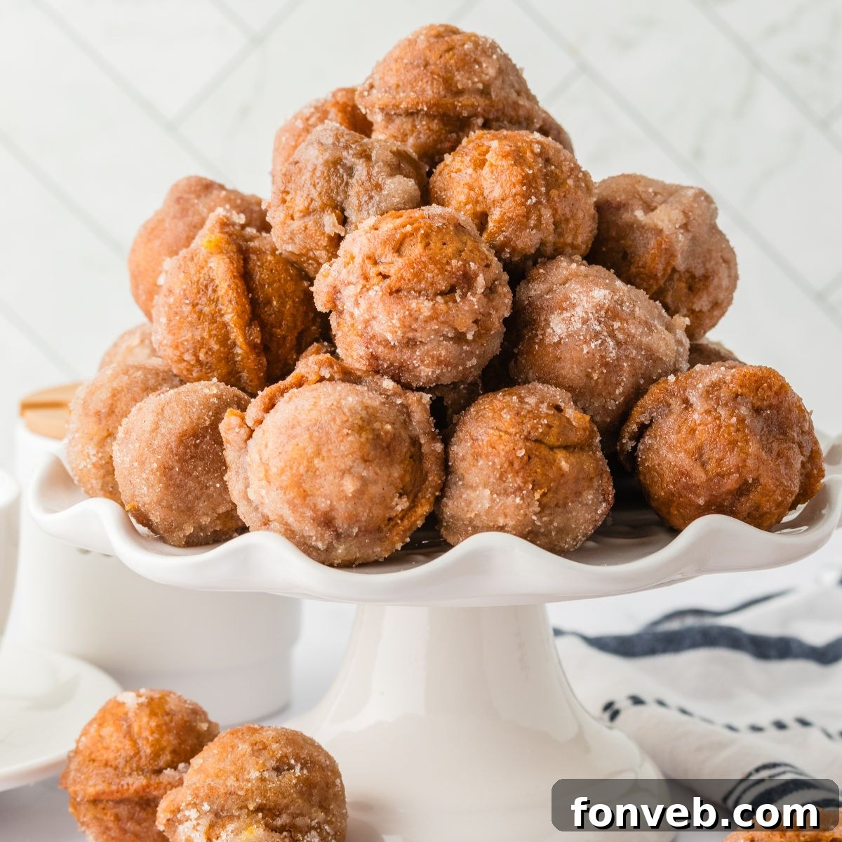 Cinnamon Sugar Baked Pumpkin Spice Donut Holes 2 Pumpkin Spice Donut Holes stacked on a cake platter sitting on table with more donut holes scattered around the table