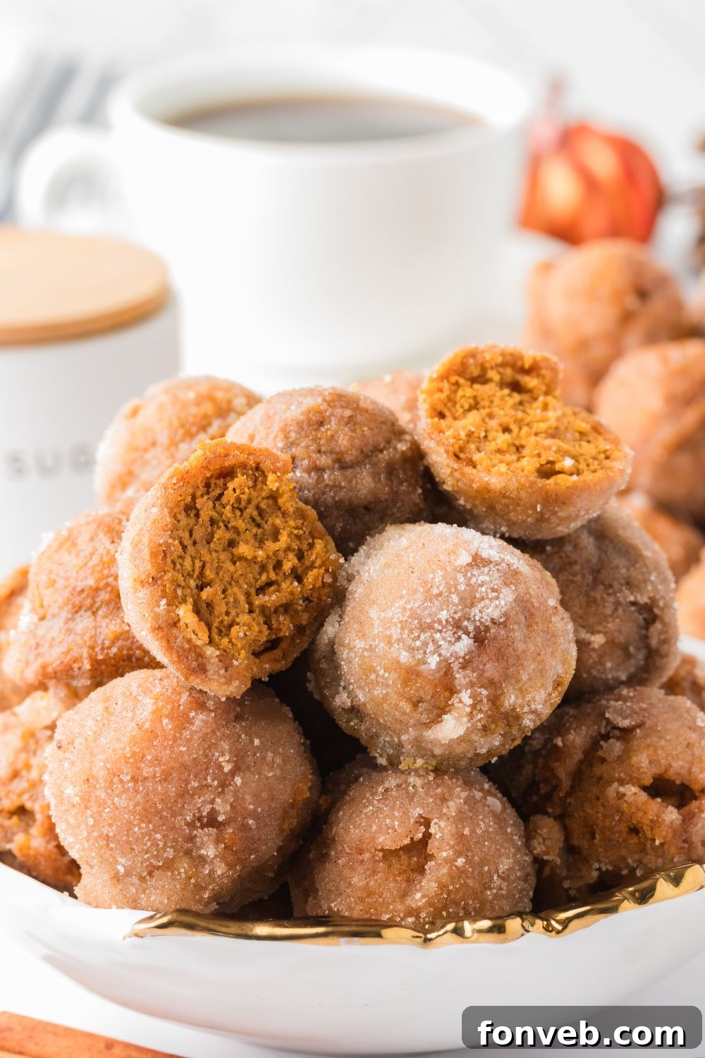 Cinnamon Sugar Baked Pumpkin Spice Donut Holes 13 Pumpkin Spice Donut Holes stacked in a bowl on the counter with a few cut in half, and a cup of coffee in the background of the image