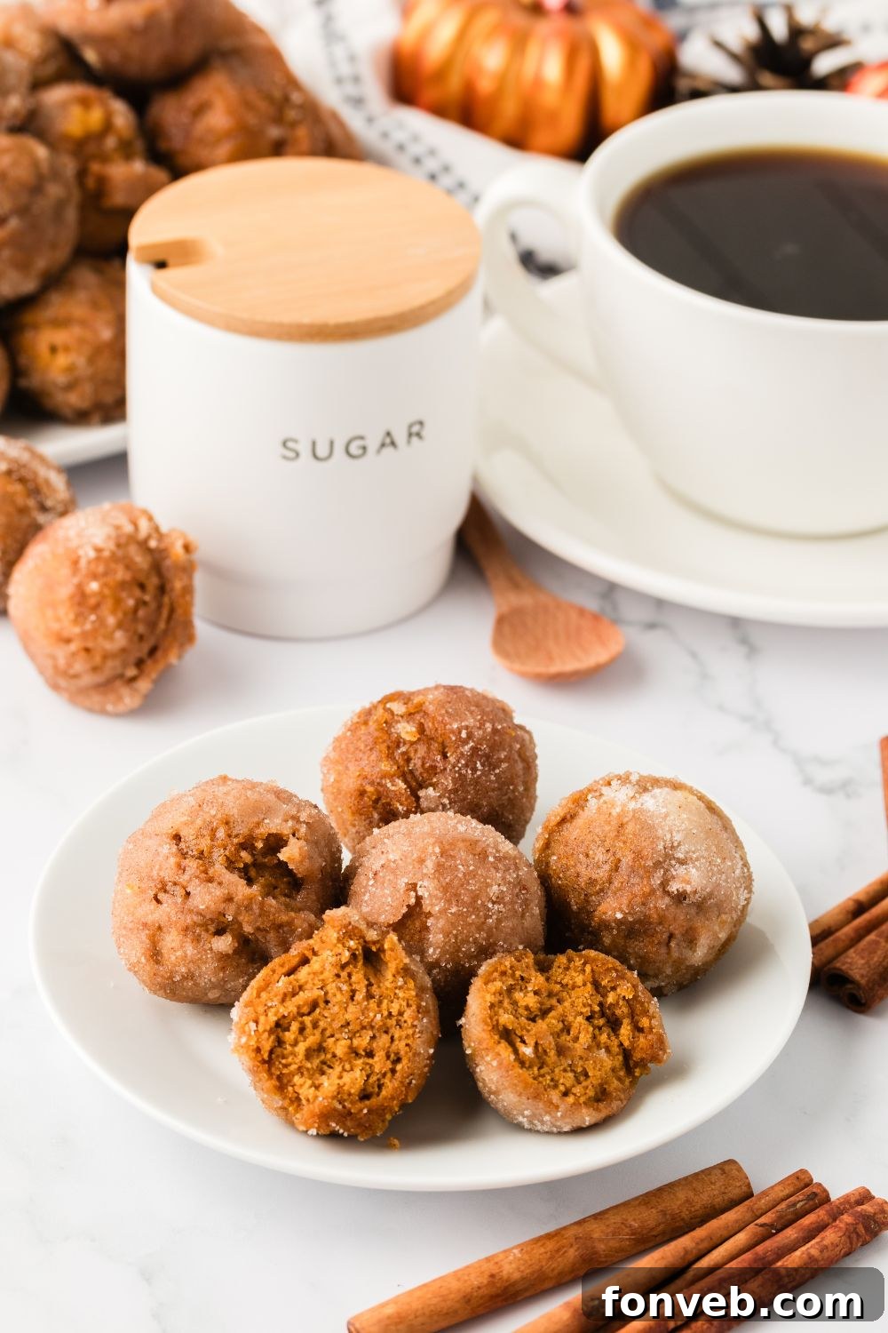 Cinnamon Sugar Baked Pumpkin Spice Donut Holes 16 plate with pumpkin spice donut holes and some are cut in half. A cup of coffee, cinnamon sticks andmore around table