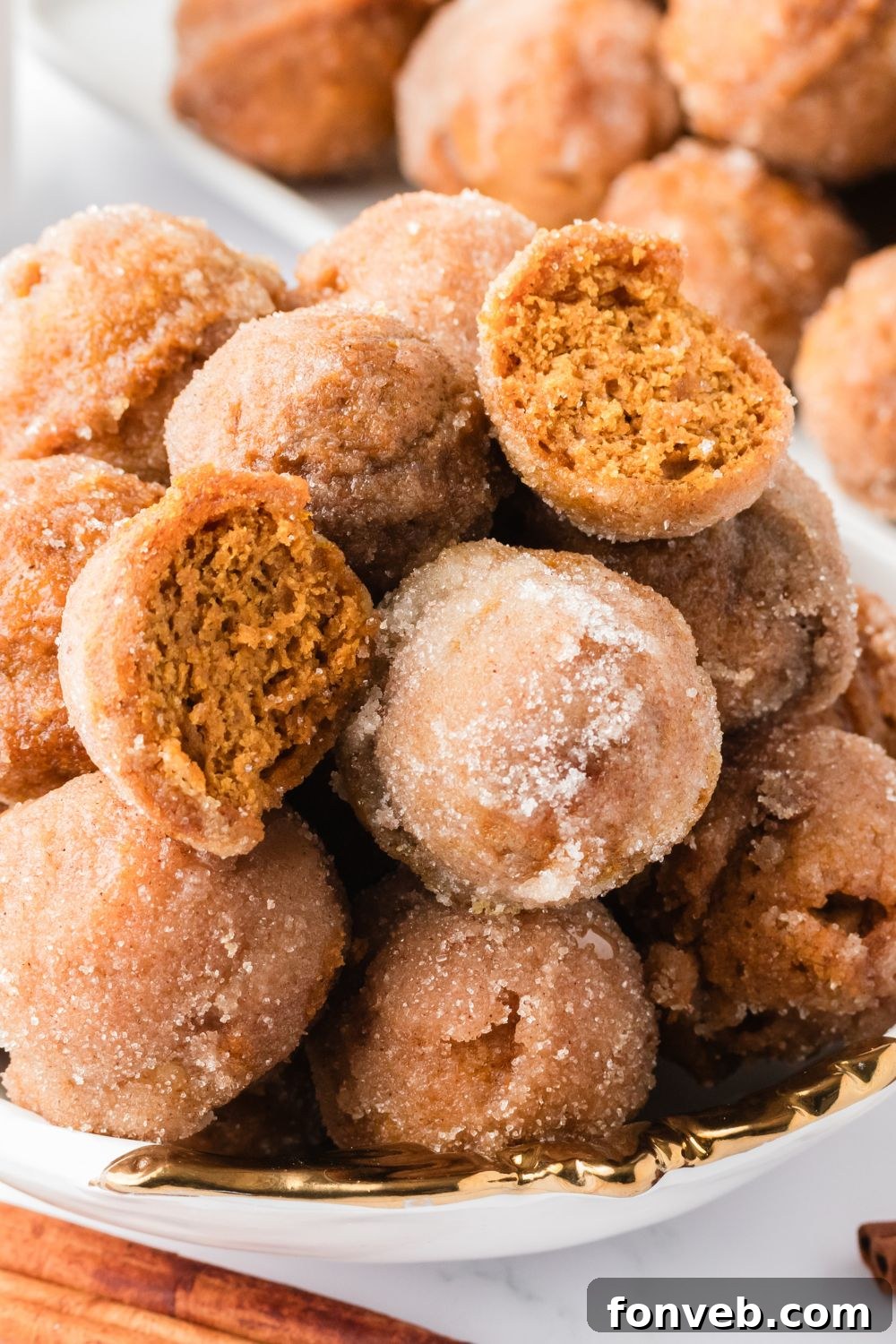 Cinnamon Sugar Baked Pumpkin Spice Donut Holes 18 Pumpkin Spice Donut Holes stacked in a bowl on table with one cut in half, and a tray full of donuts behind it on table