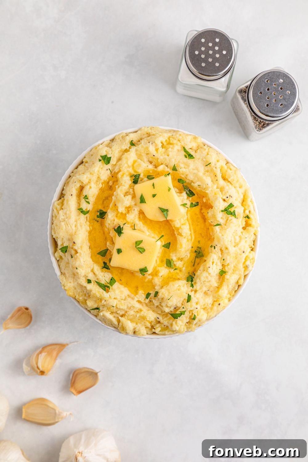 Effortless Creamy Garlic Mashed Potatoes 11 large white bowl on table full of Garlic Mashed Potatoes and garlic to side and salt and pepper shakers