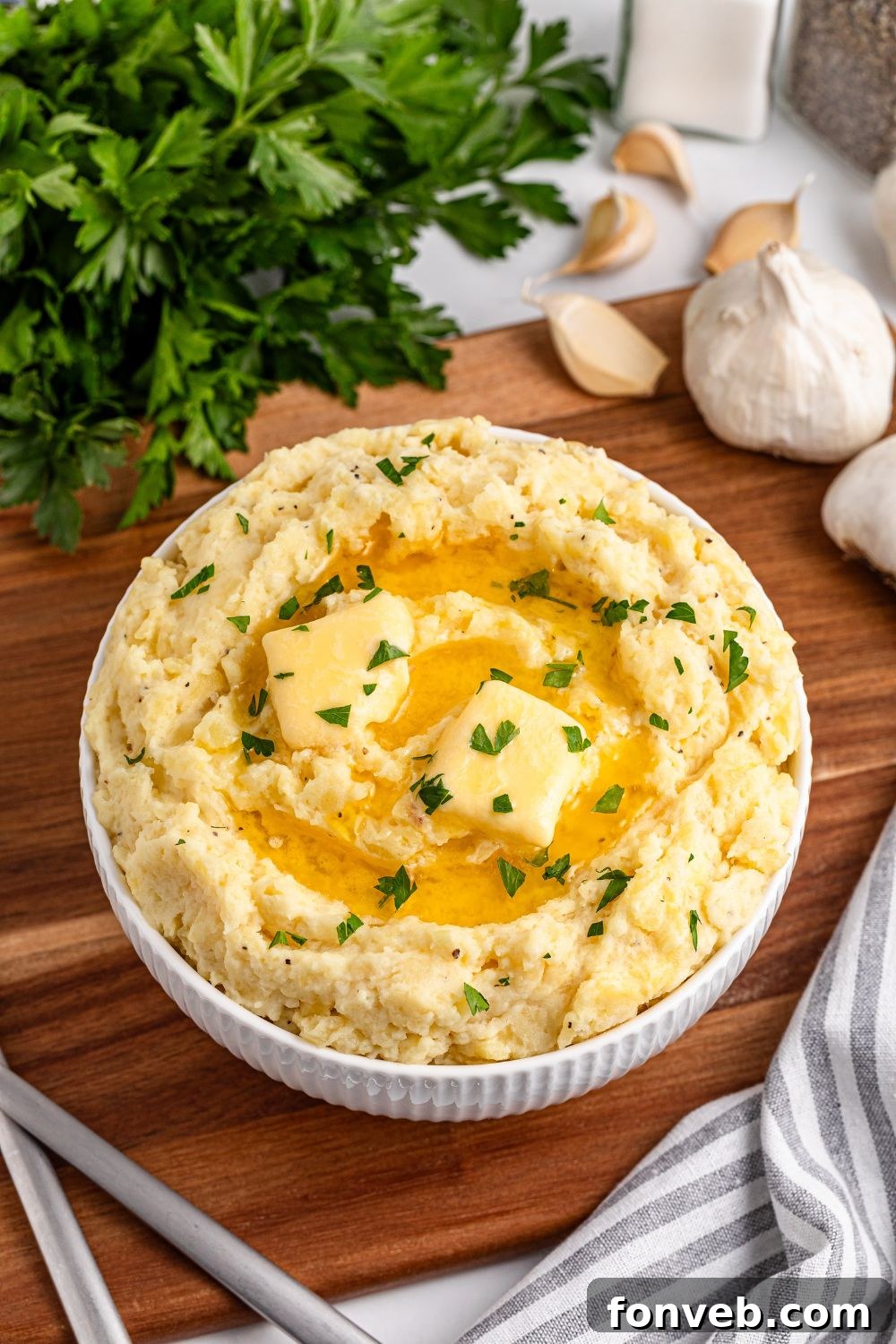 Effortless Creamy Garlic Mashed Potatoes 12 a wooden cutting board with a white bowl full of Garlic Mashed Potatoes in it, topped with melted butter and parsley. To the side is fresh garlic, a towel, and parsley
