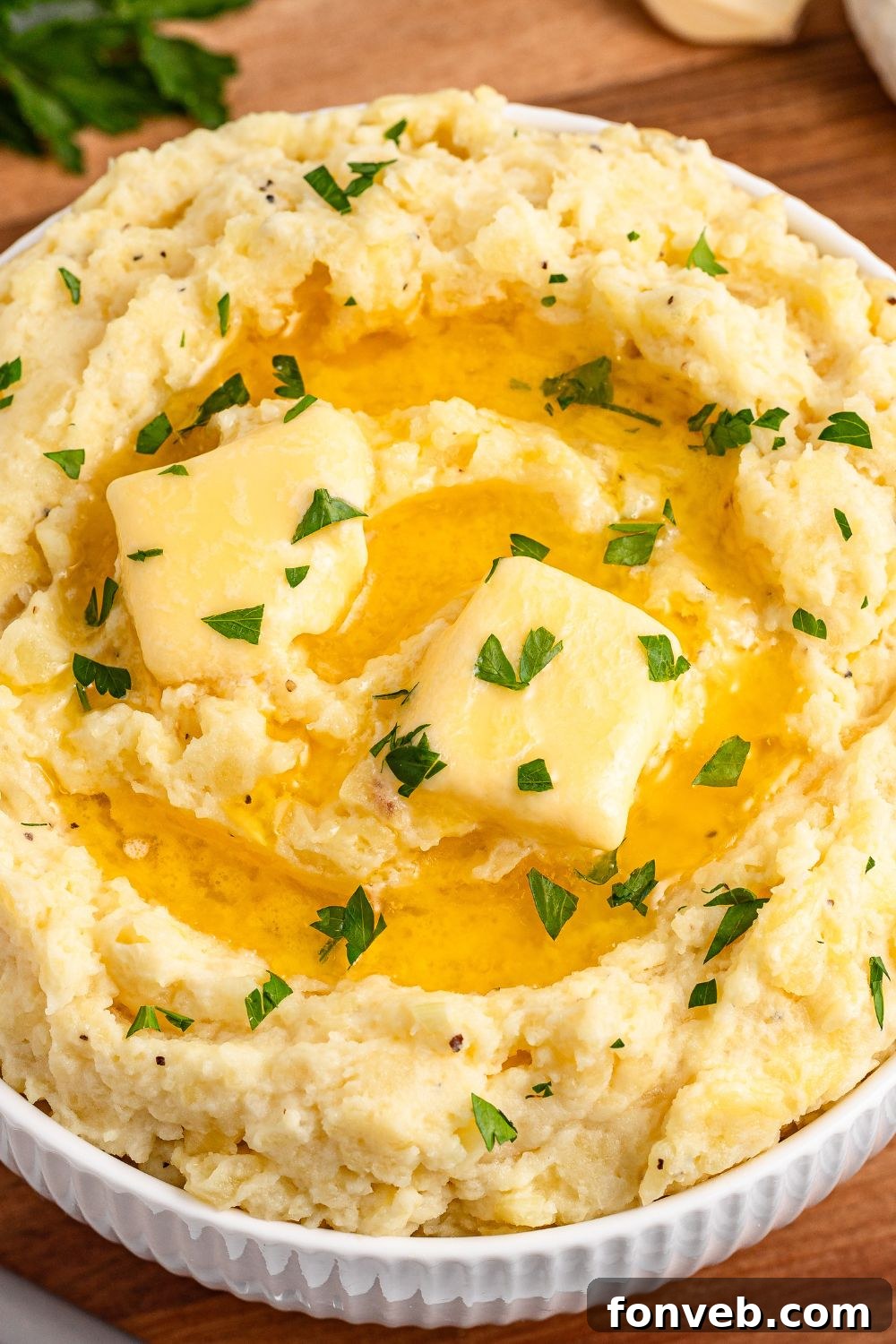 Effortless Creamy Garlic Mashed Potatoes 13 close up look of Garlic Mashed Potatoes in a white bowl on wooden cutting board. The potatoes are topped with melted butter and fresh parsley