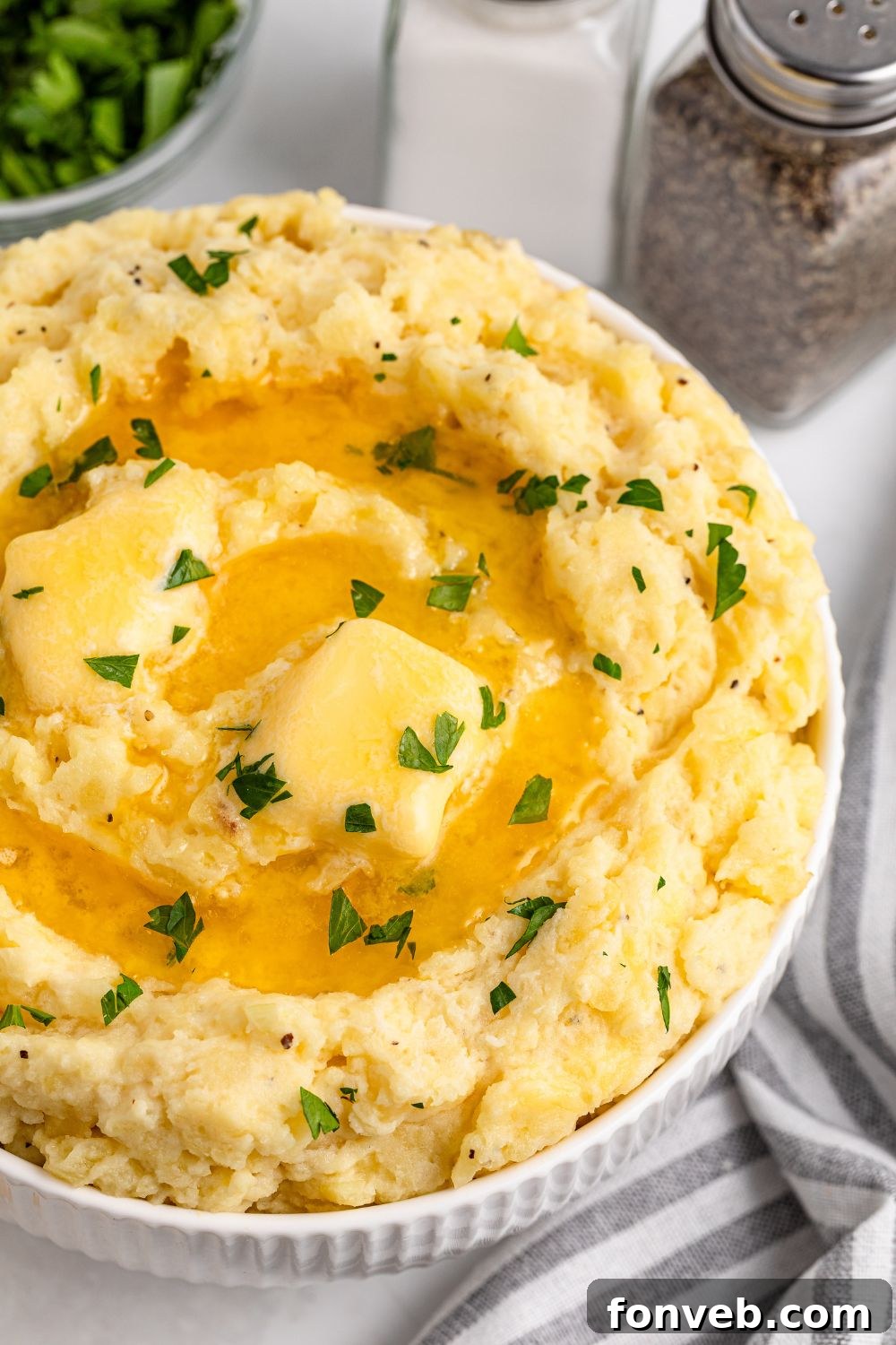 Effortless Creamy Garlic Mashed Potatoes 16 view of part of a white bowl on table full of Garlic Mashed Potatoes with melted butter and chopped parsley on top. A salt and pepper shaker are to the side of the bowl