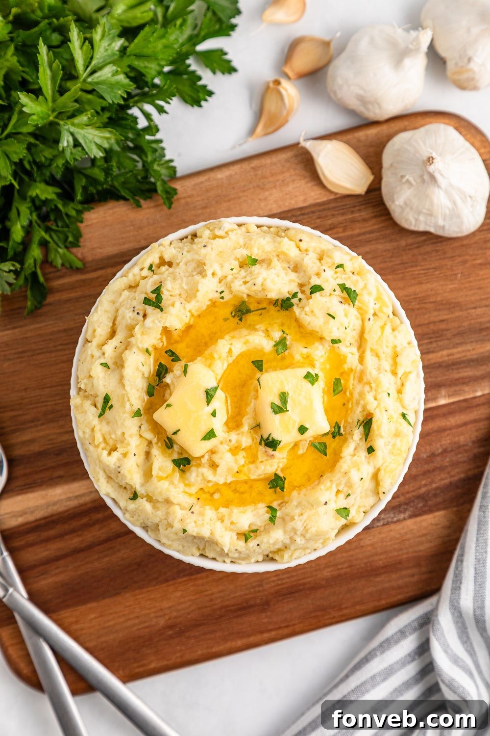 Effortless Creamy Garlic Mashed Potatoes 18 Looking down at table to see a wooden cutting board with a bowl of Garlic Mashed Potatoes in the center. To the side a bundle of parsley and fresh garlic cloves
